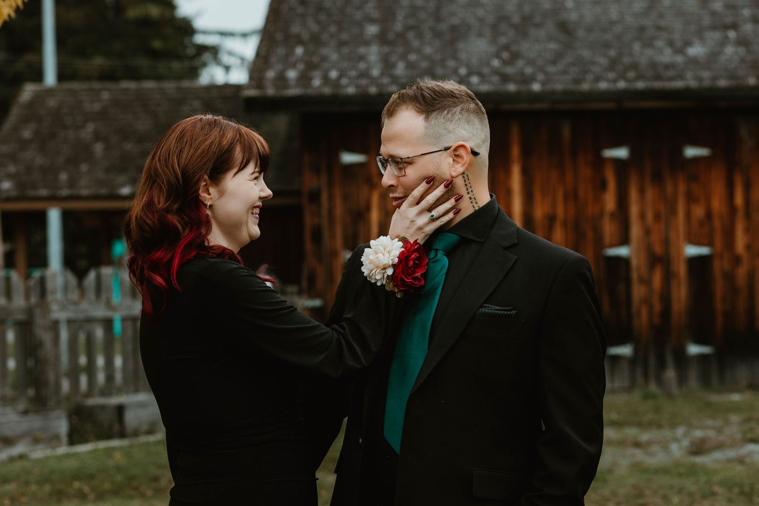 Two people share an intimate moment in formal attire outside a rustic barn building.