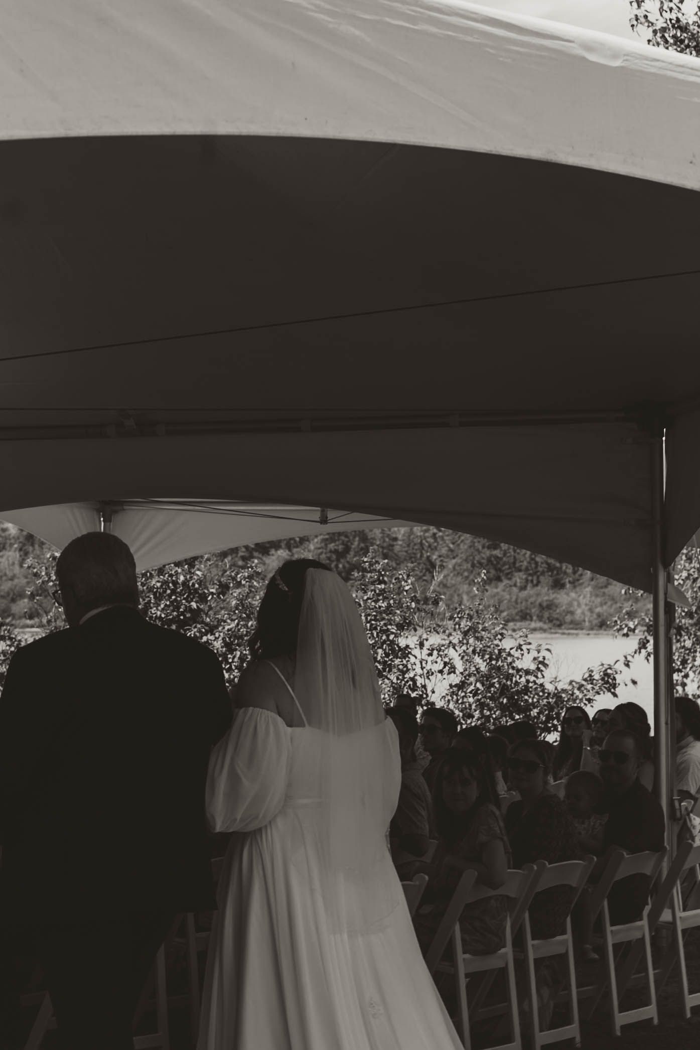 A wedding ceremony takes place under a white tent with guests seated on wooden folding chairs.