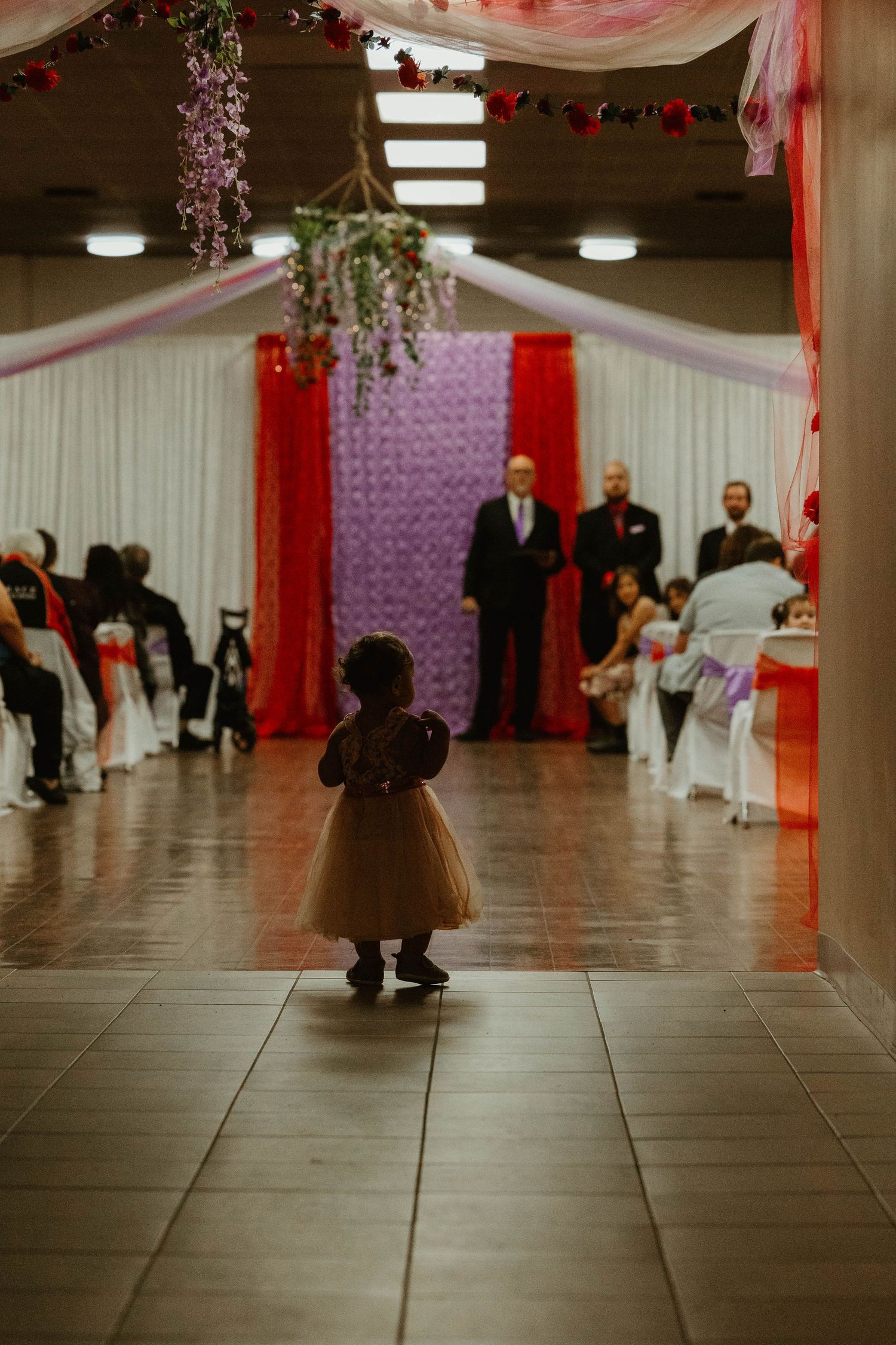 Small child in brown dress stands on dance floor with red and purple wedding decorations.