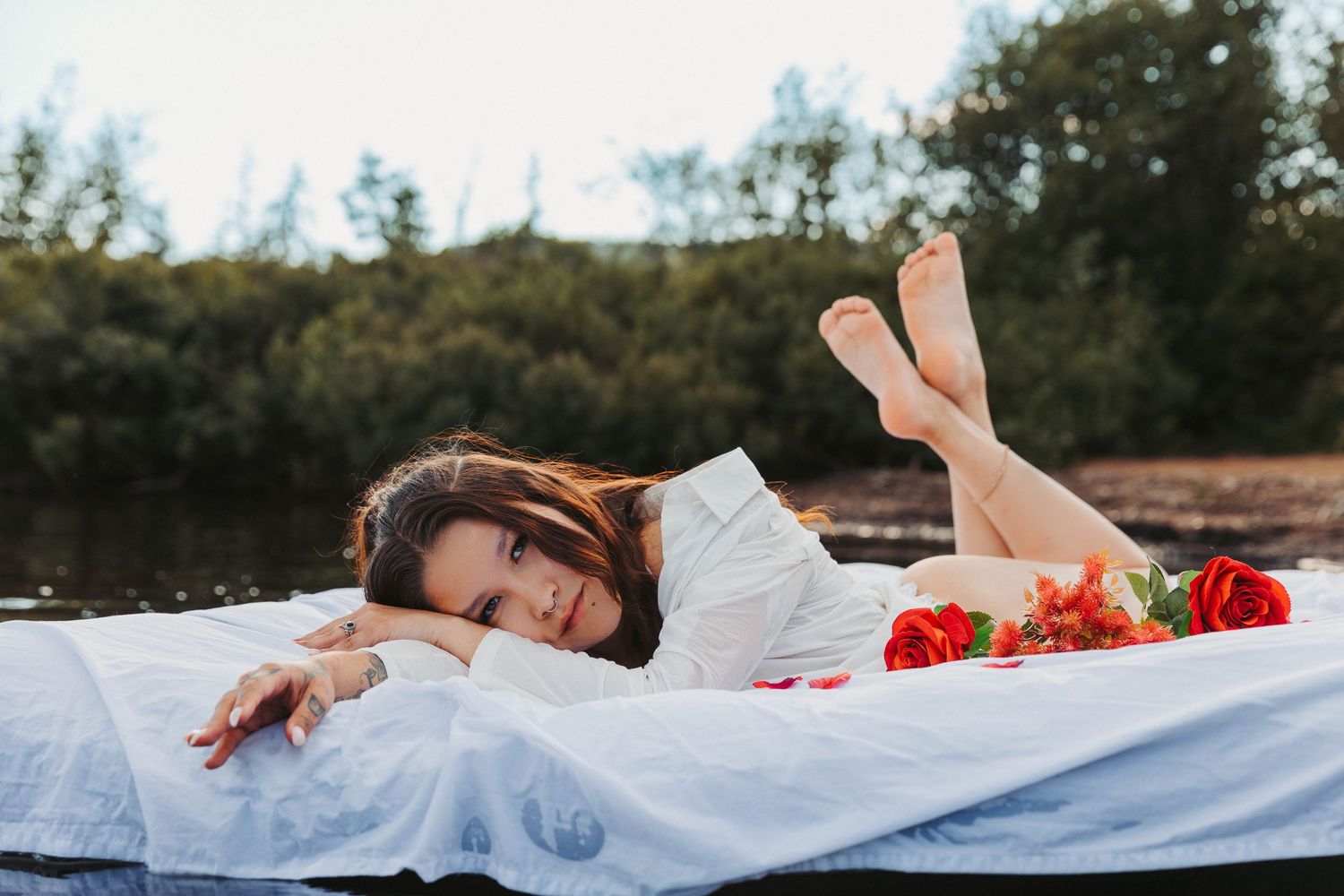 Person in white dress laying on blanket outdoors with red flowers scattered around.