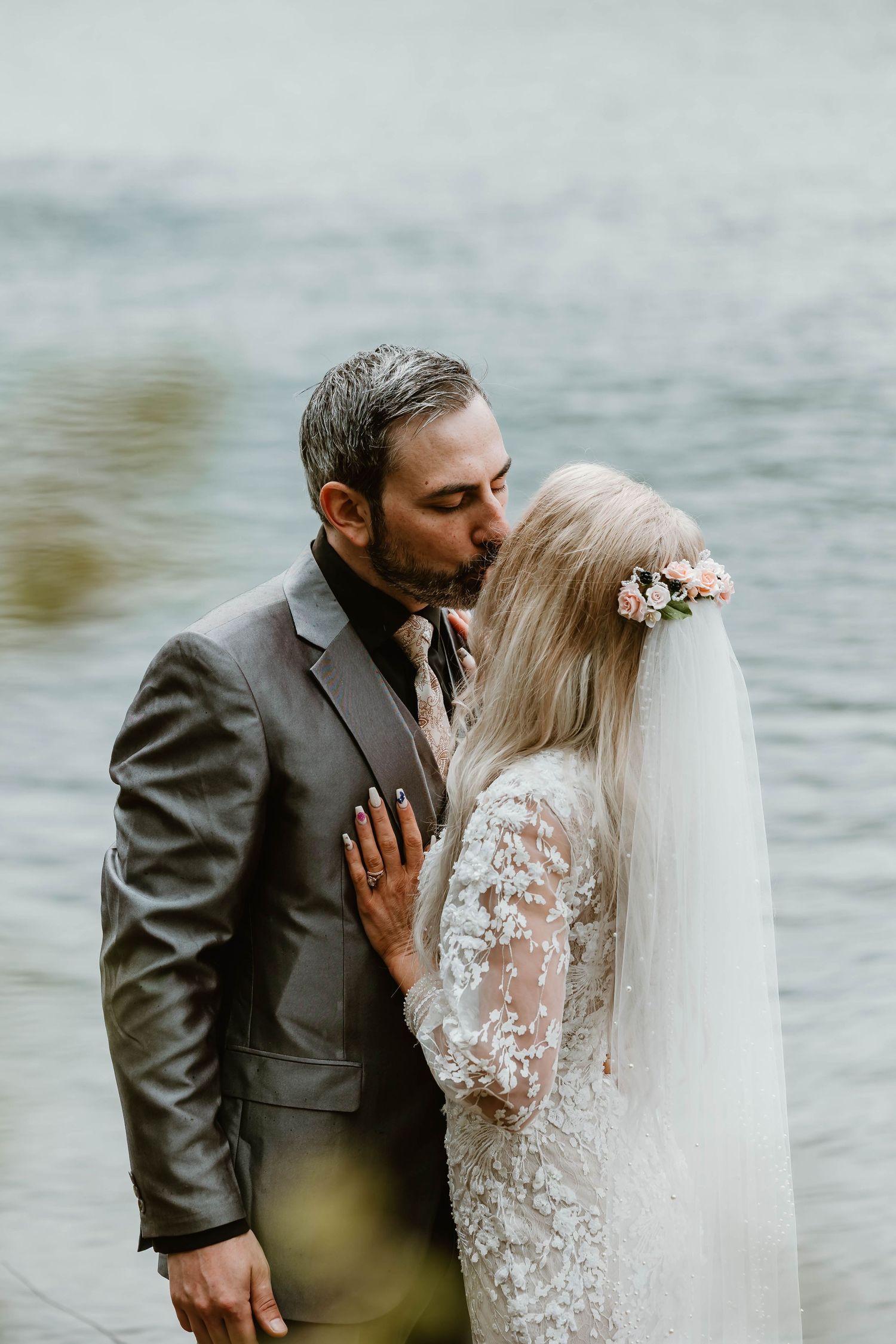 A romantic wedding couple embraces by the water, with the bride wearing a lace gown and floral headpiece with flowing veil.