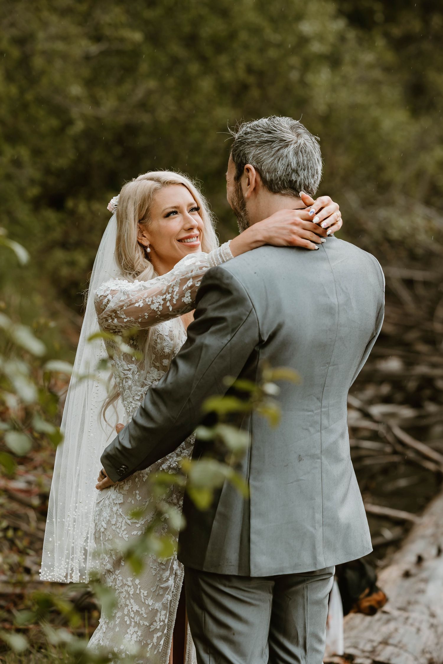 A romantic wedding moment captured in nature showing a couple in grey suit and delicate lace dress embracing outdoors.