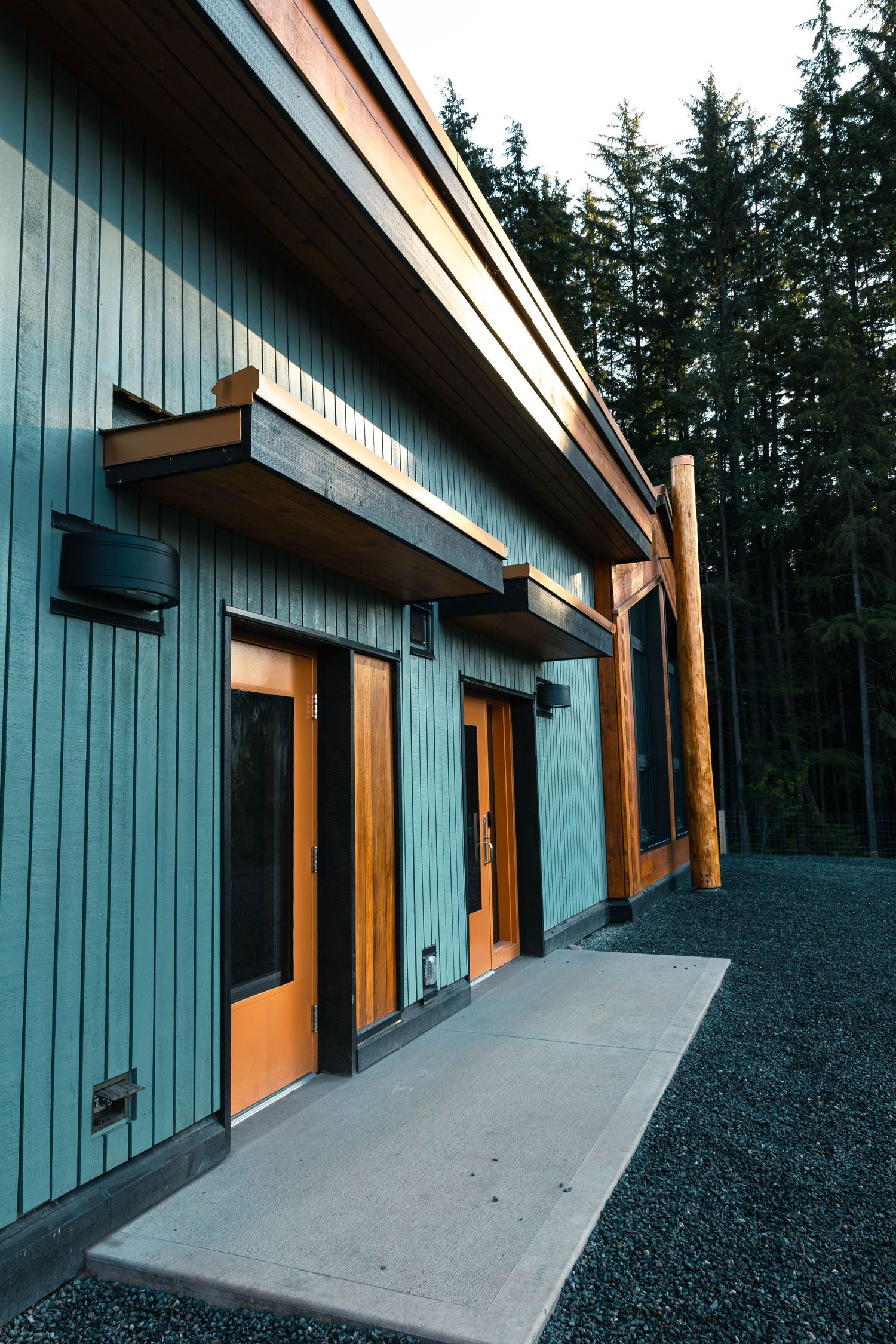 Modern rustic building exterior with teal vertical siding, orange doors, wooden beams and gravel path against evergreen forest.
