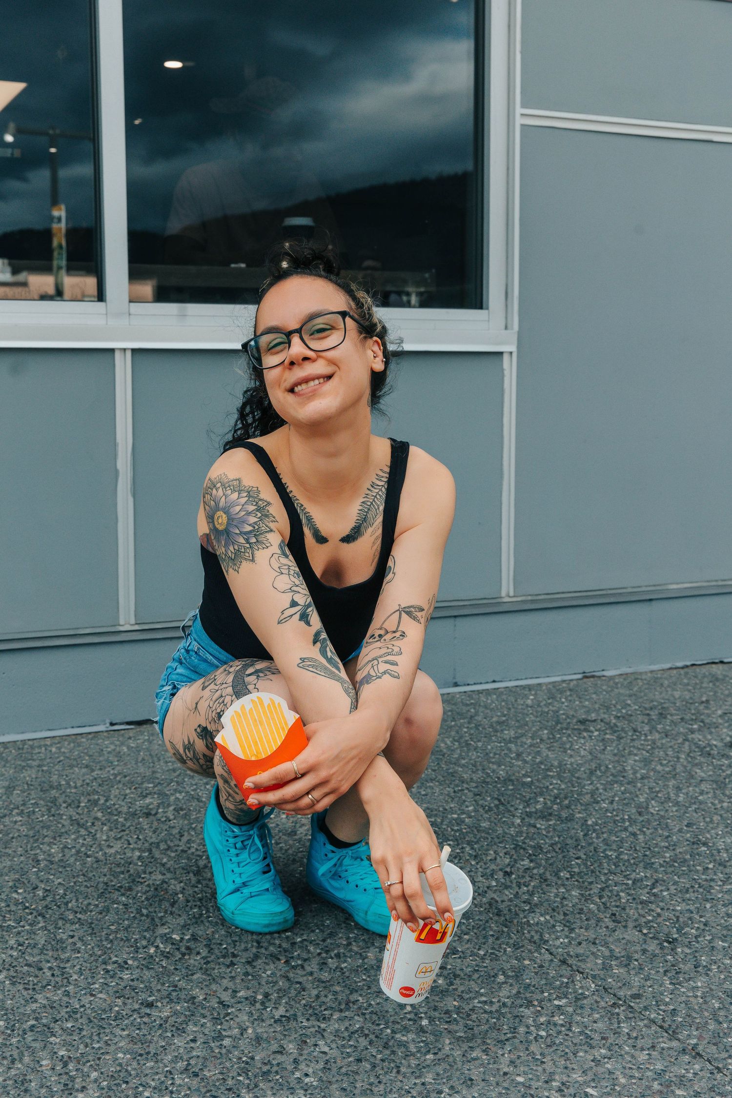Casual urban portrait with tattoos and bright blue shoes outside a storefront.