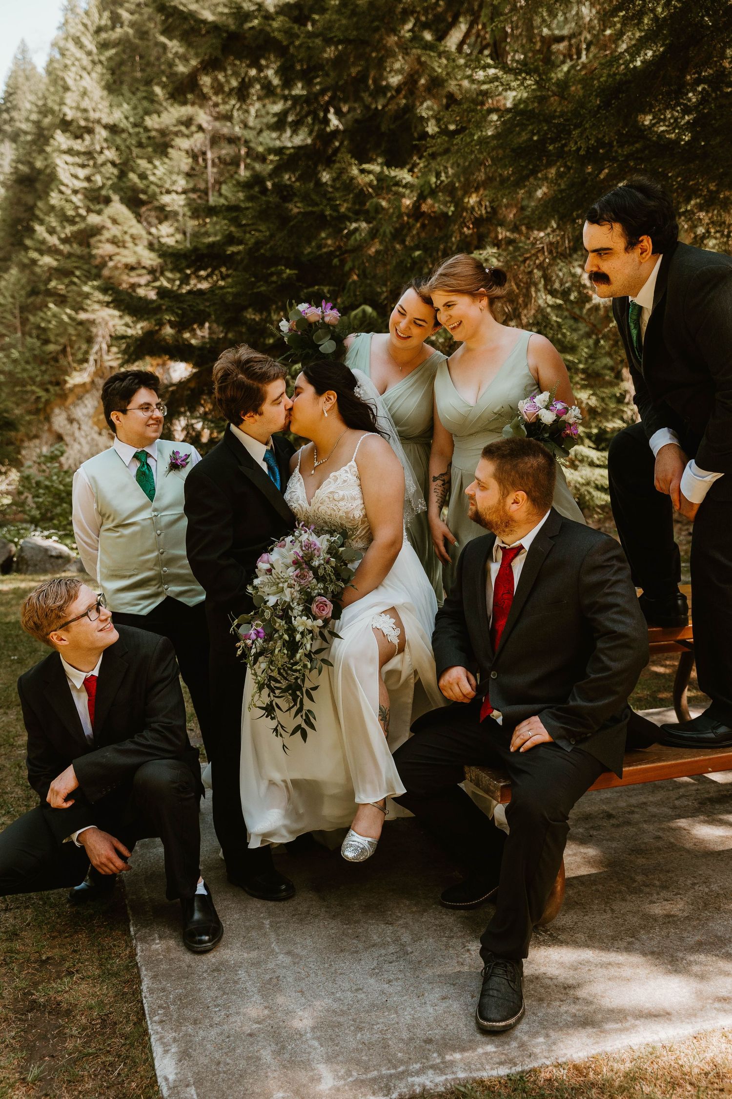 Wedding party gathered for casual group photo in forest with pine trees in background.