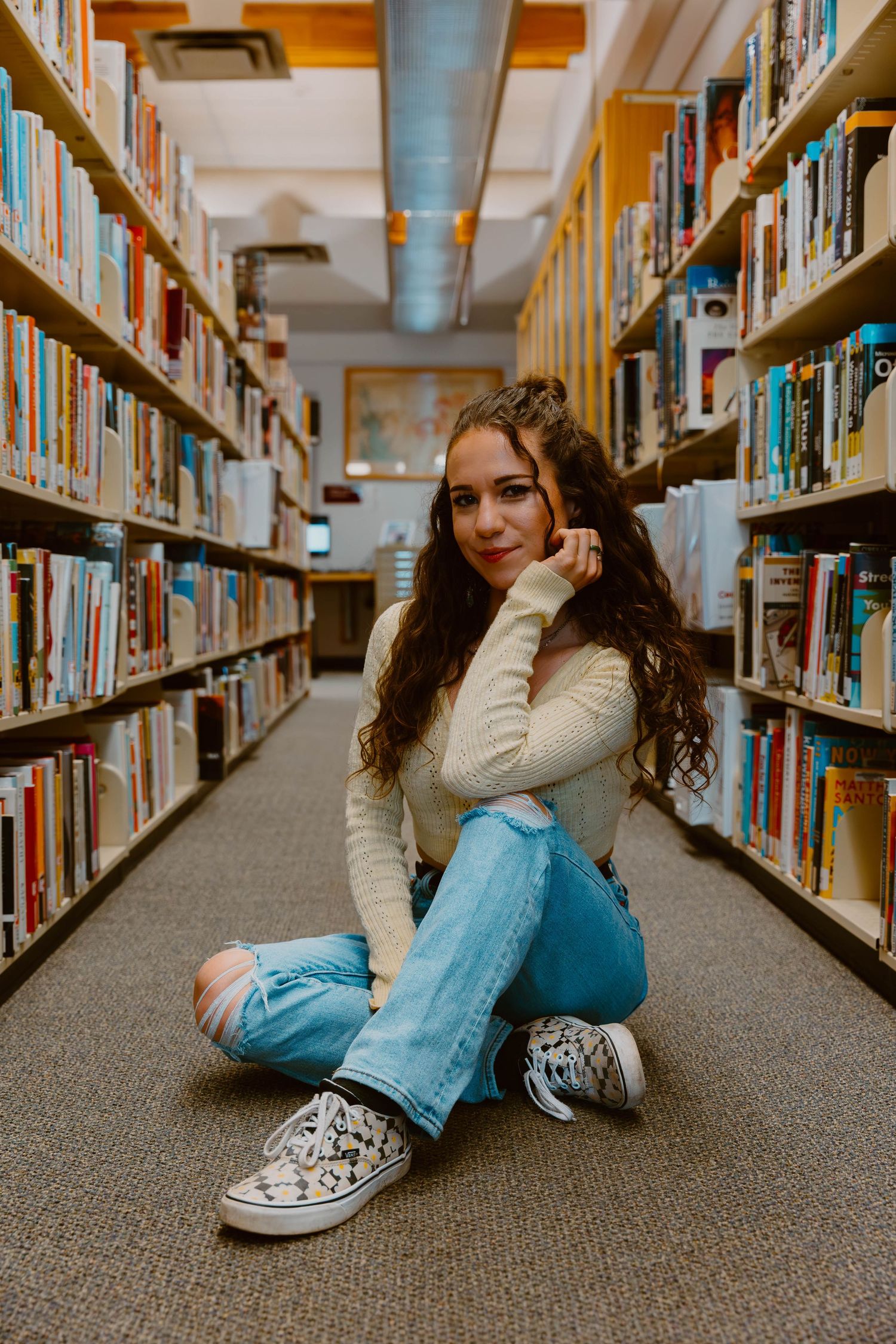 Student sits cross-legged between library bookshelves wearing casual attire.