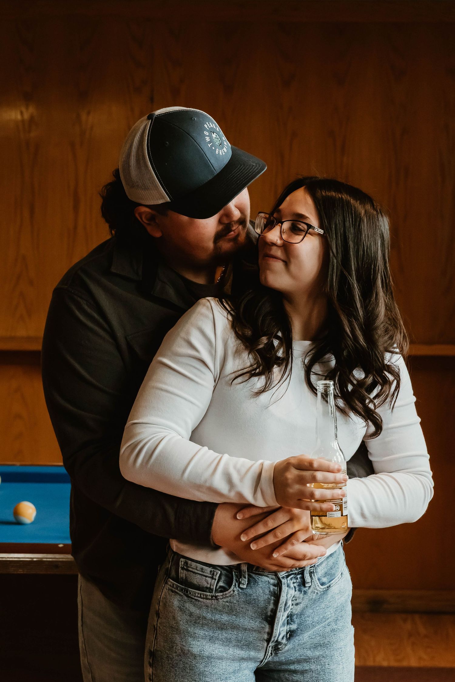 Couple shares intimate moment while holding whiskey glass against wooden wall with blue accent.