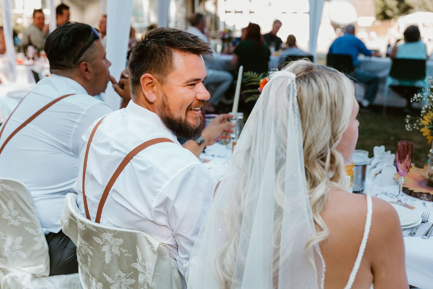 Guests gather at outdoor wedding reception tables under white tent with decorative lighting.