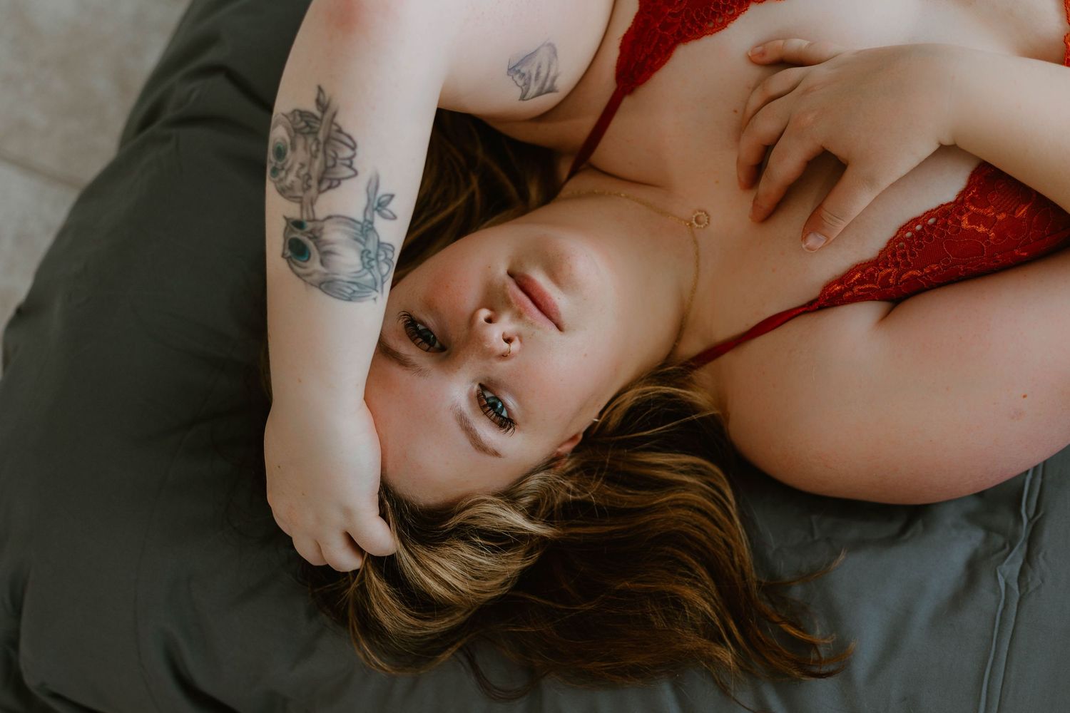 Intimate boudoir portrait with red lingerie and visible arm tattoos against dark bedding.