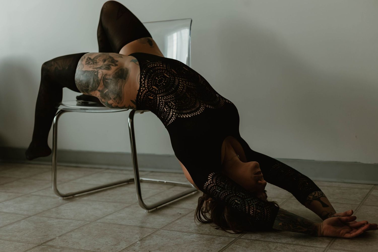 A figure in black lace reclines dramatically across a modern chair in a moody indoor setting.