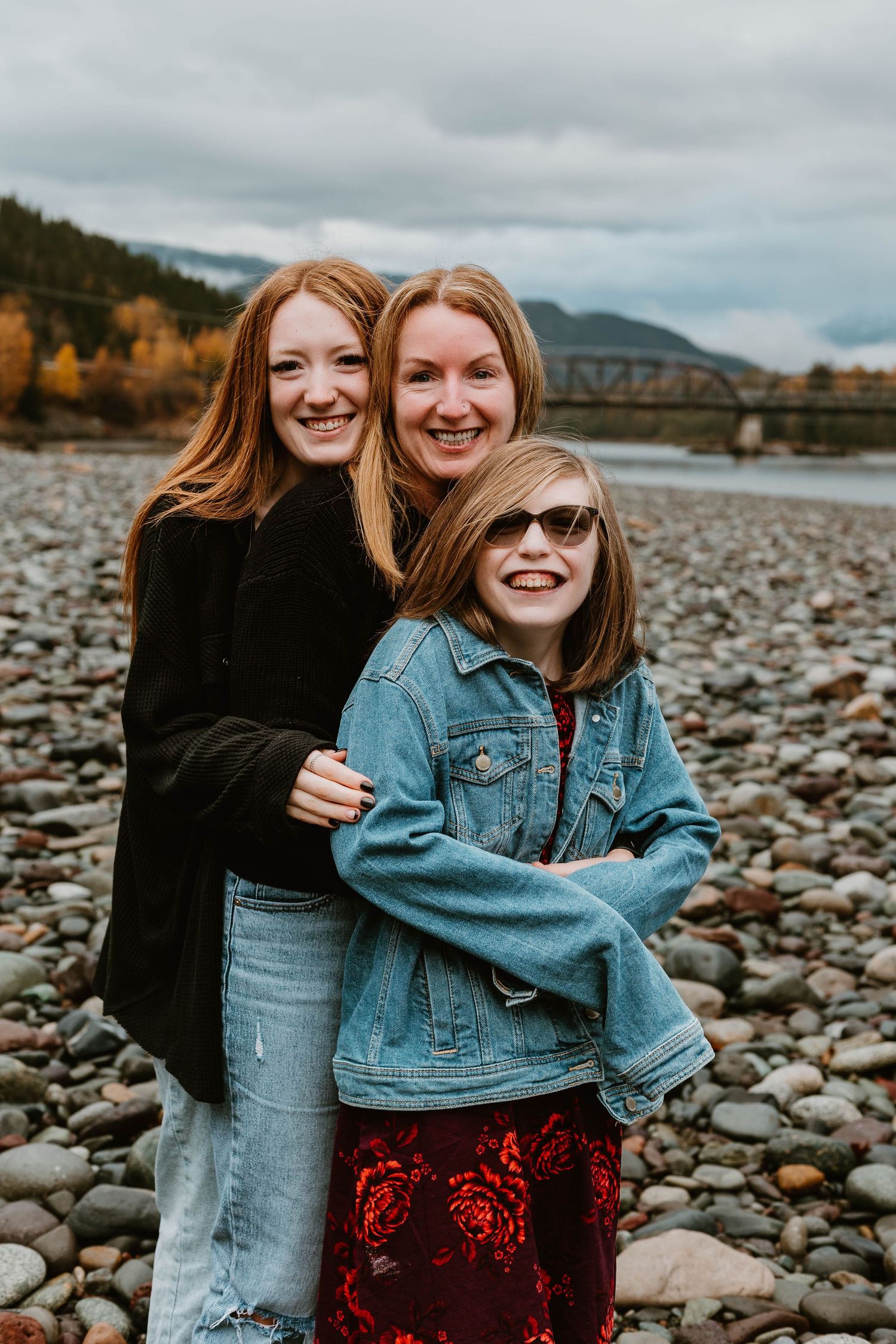 Three people posing together on rocky riverbank with mountains and bridge in background.