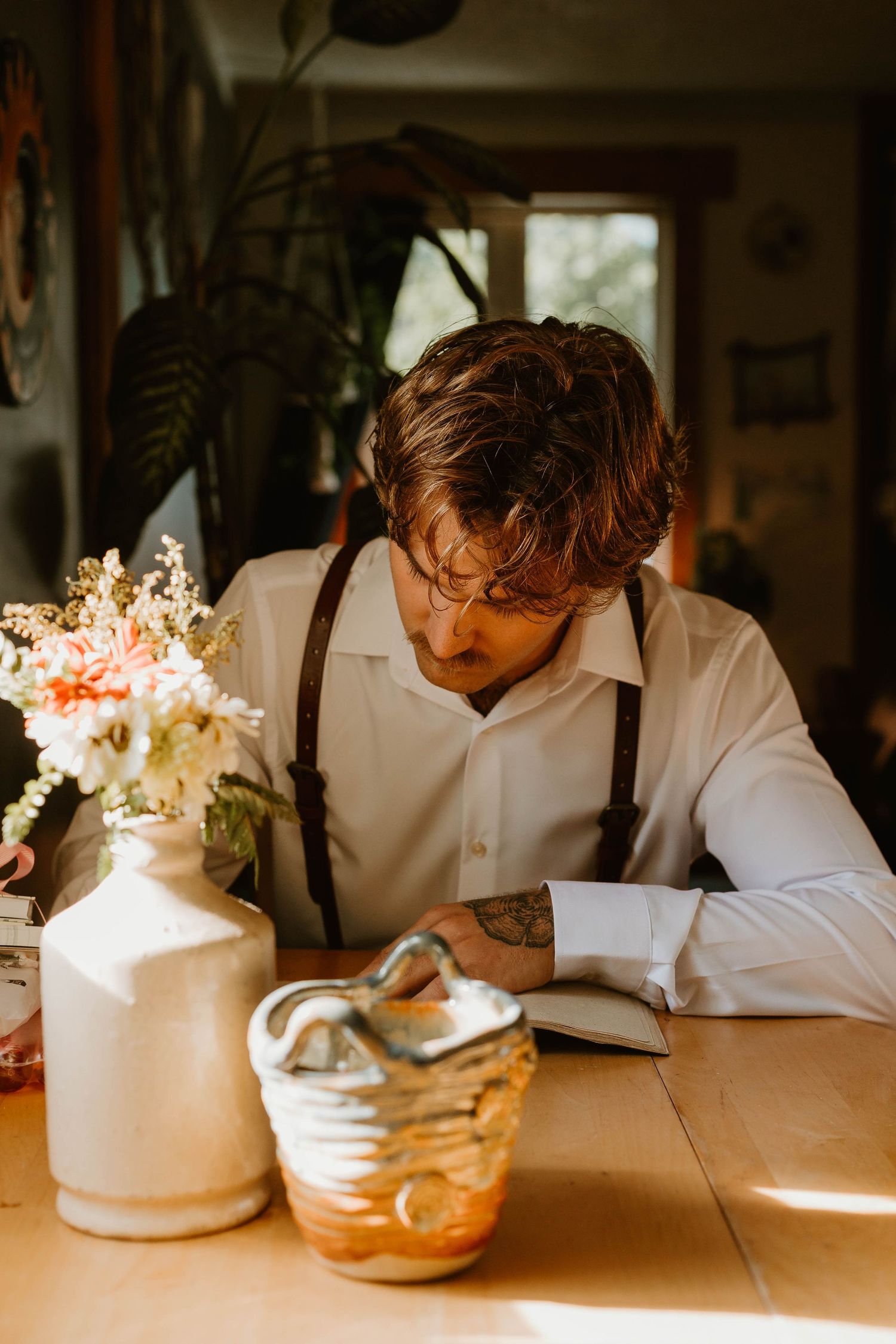 Person in white shirt and suspenders sits at table with flowers and notebook.