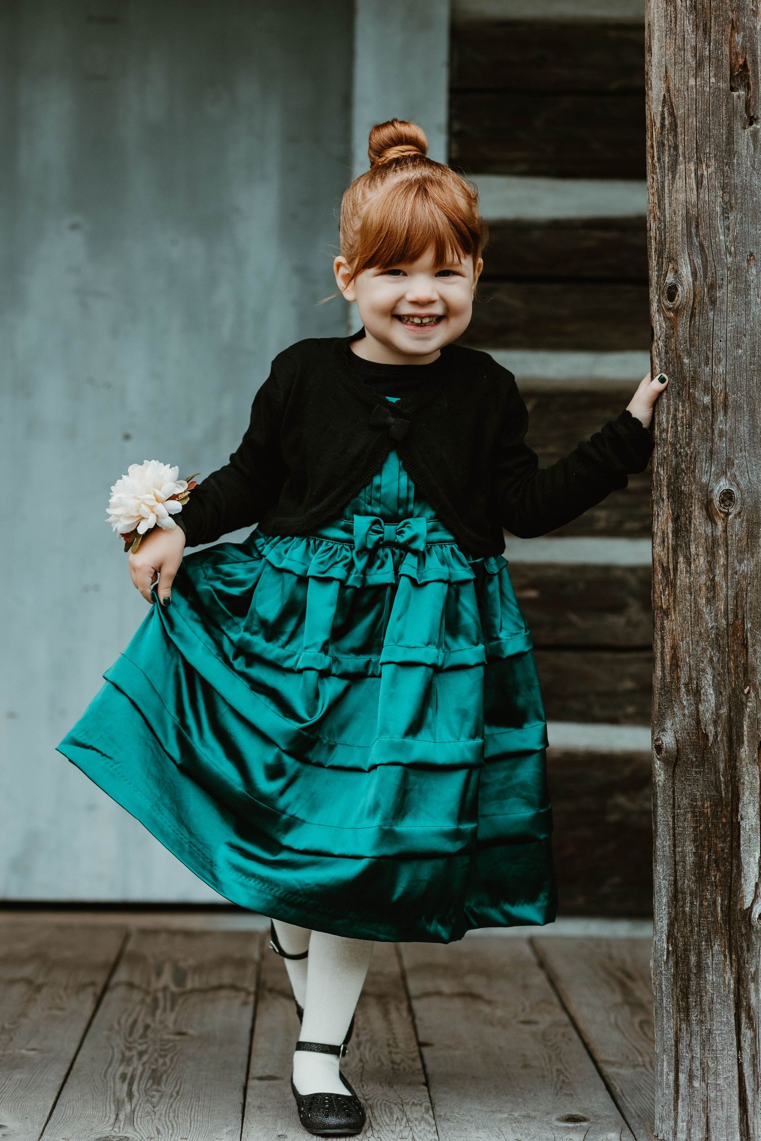 A young child in a teal and black party dress poses playfully while holding a flower on a wooden porch.
