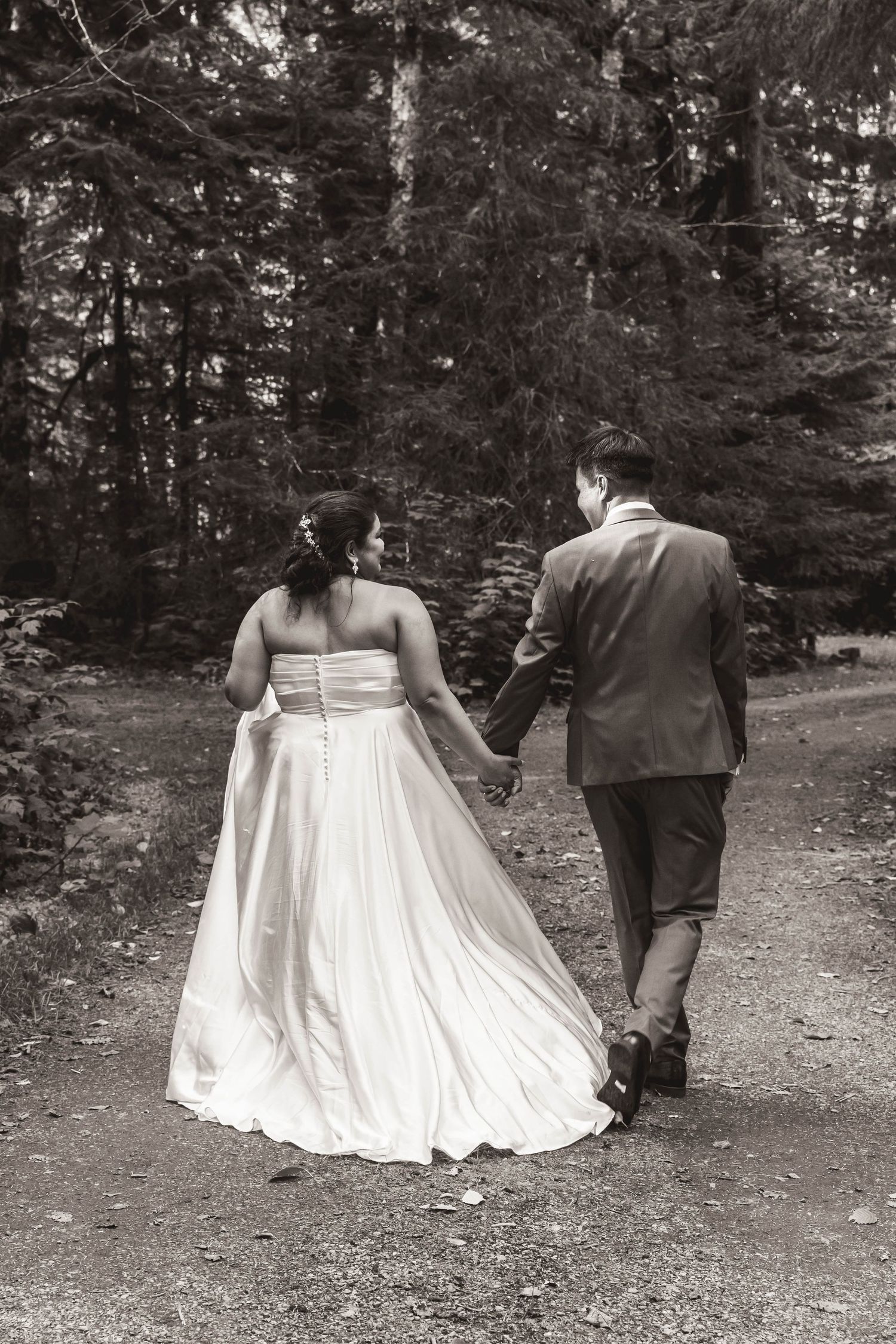Black and white photo of newlyweds walking hand in hand down a forest trail.