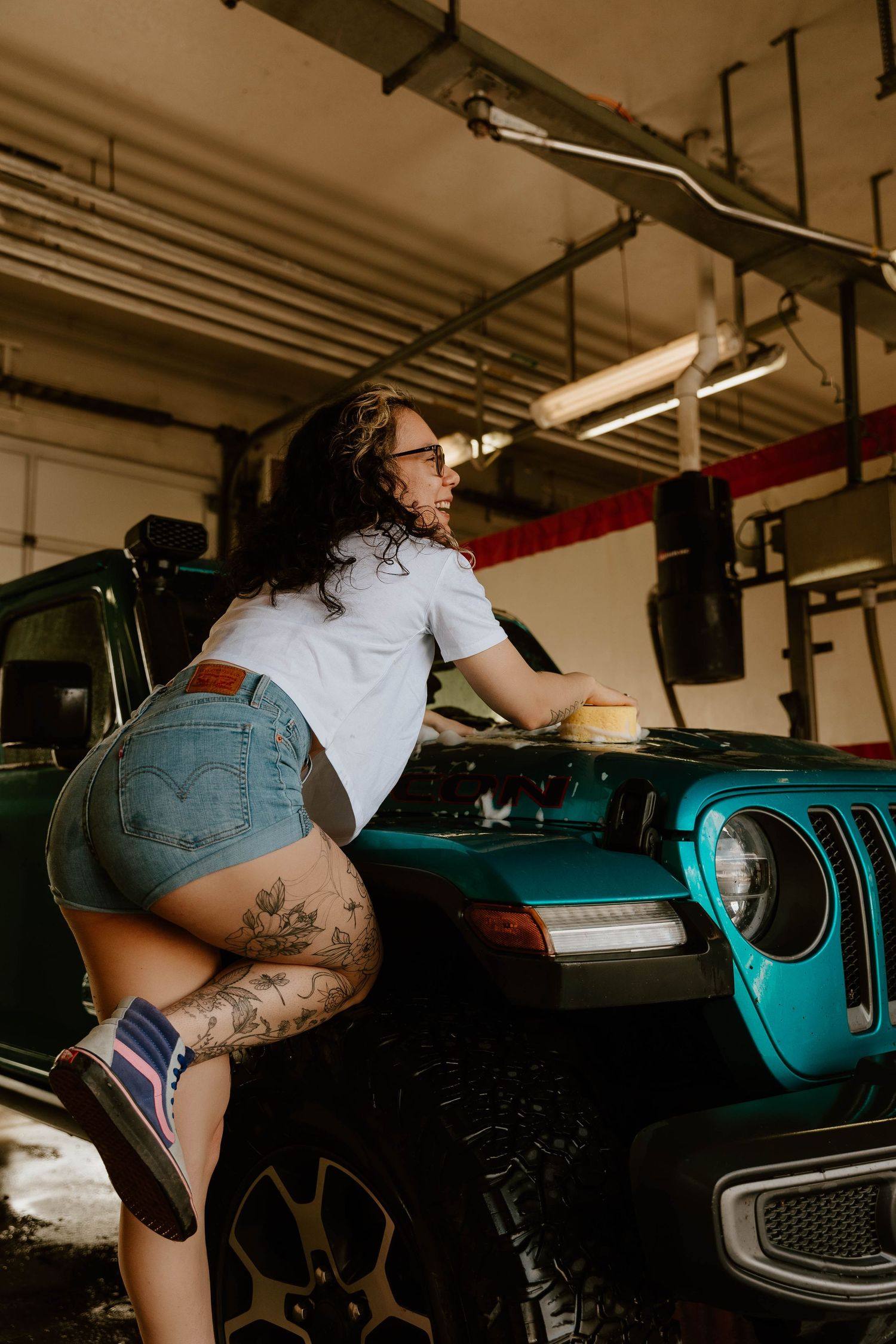Person in denim shorts and white shirt poses with vintage teal Jeep in industrial garage setting.
