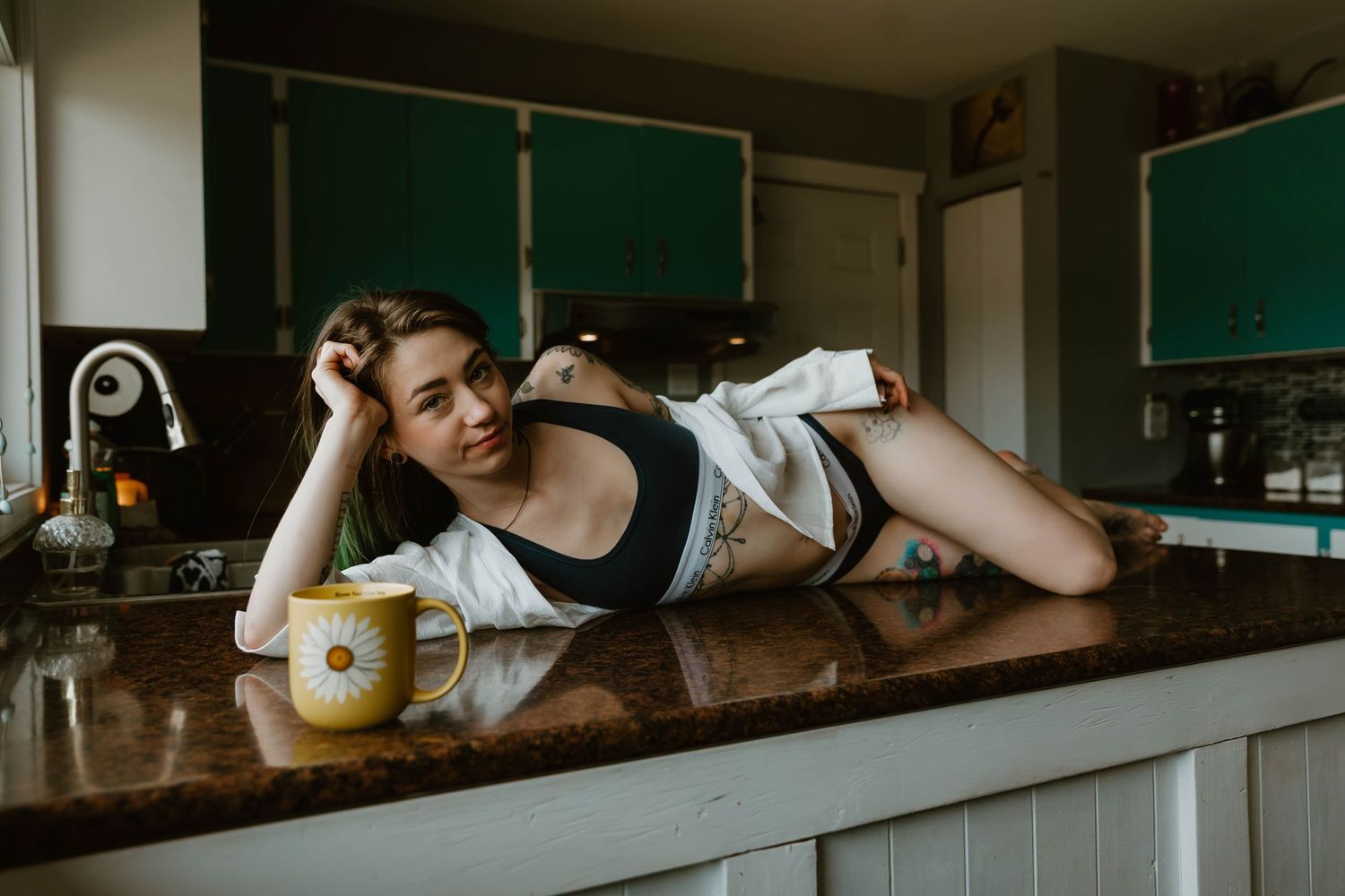 A person in black athletic wear lies on a wooden kitchen counter next to a yellow mug in moody lighting.