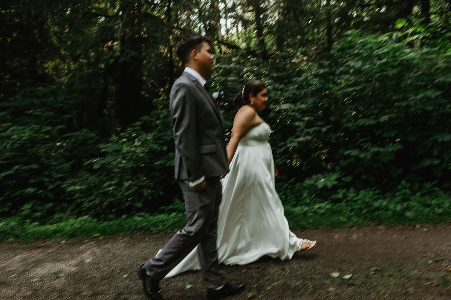 A couple in wedding attire walking through a forest path.