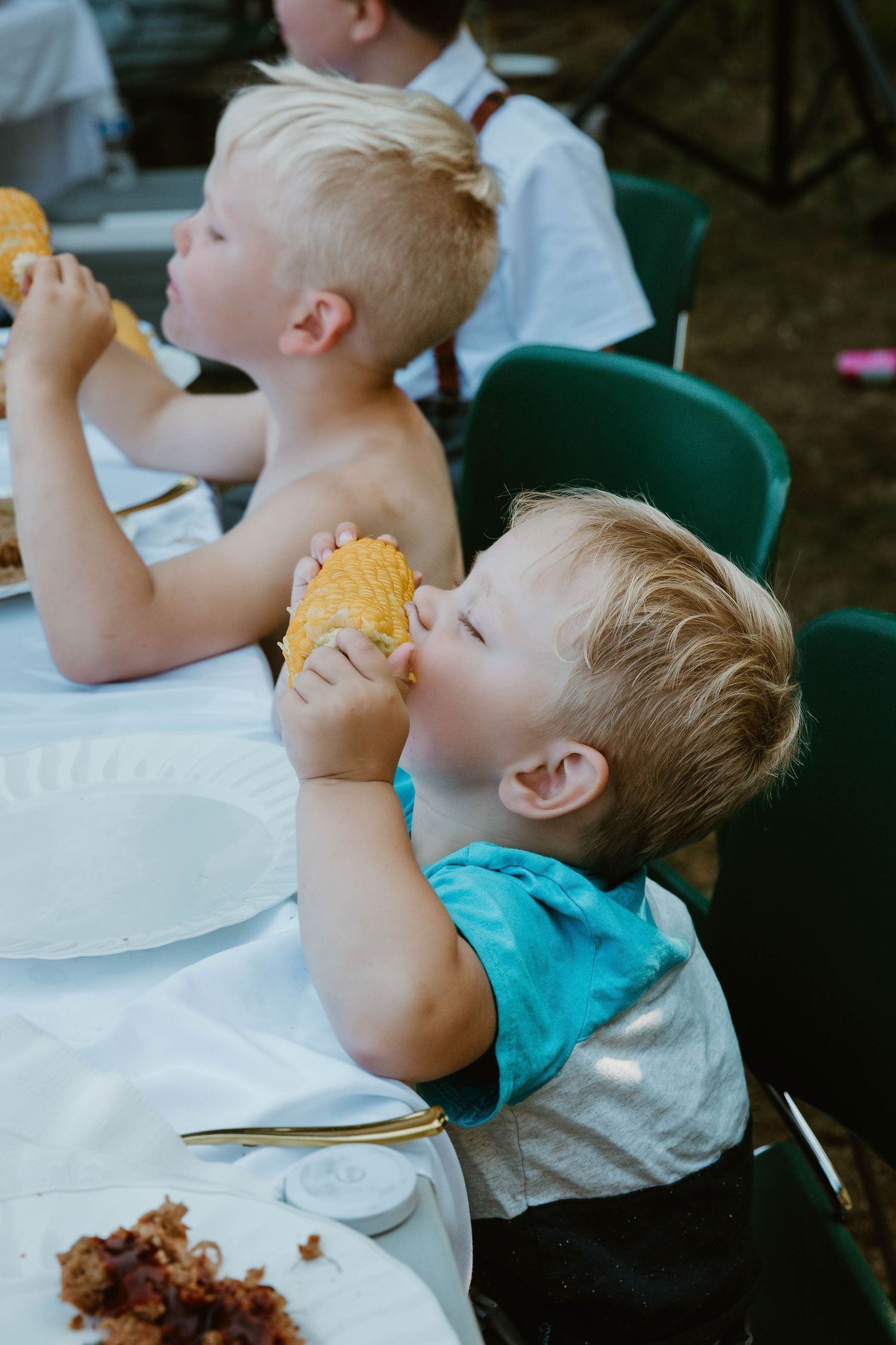 Two young boys in casual attire eagerly eating corn on the cob at an outdoor event.