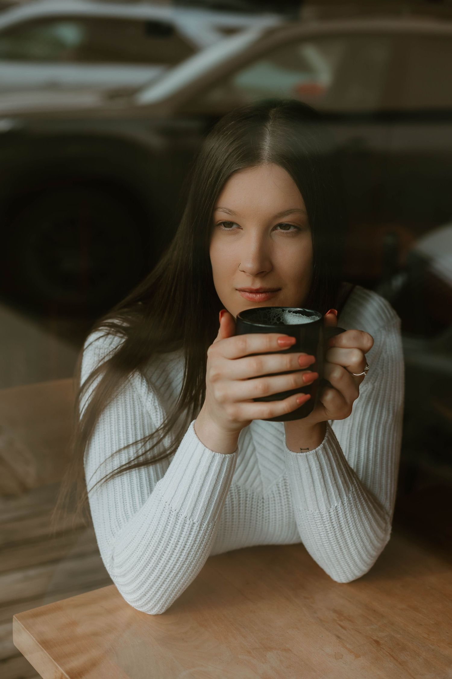 Person in white sweater holding dark coffee cup while sitting at wooden table.
