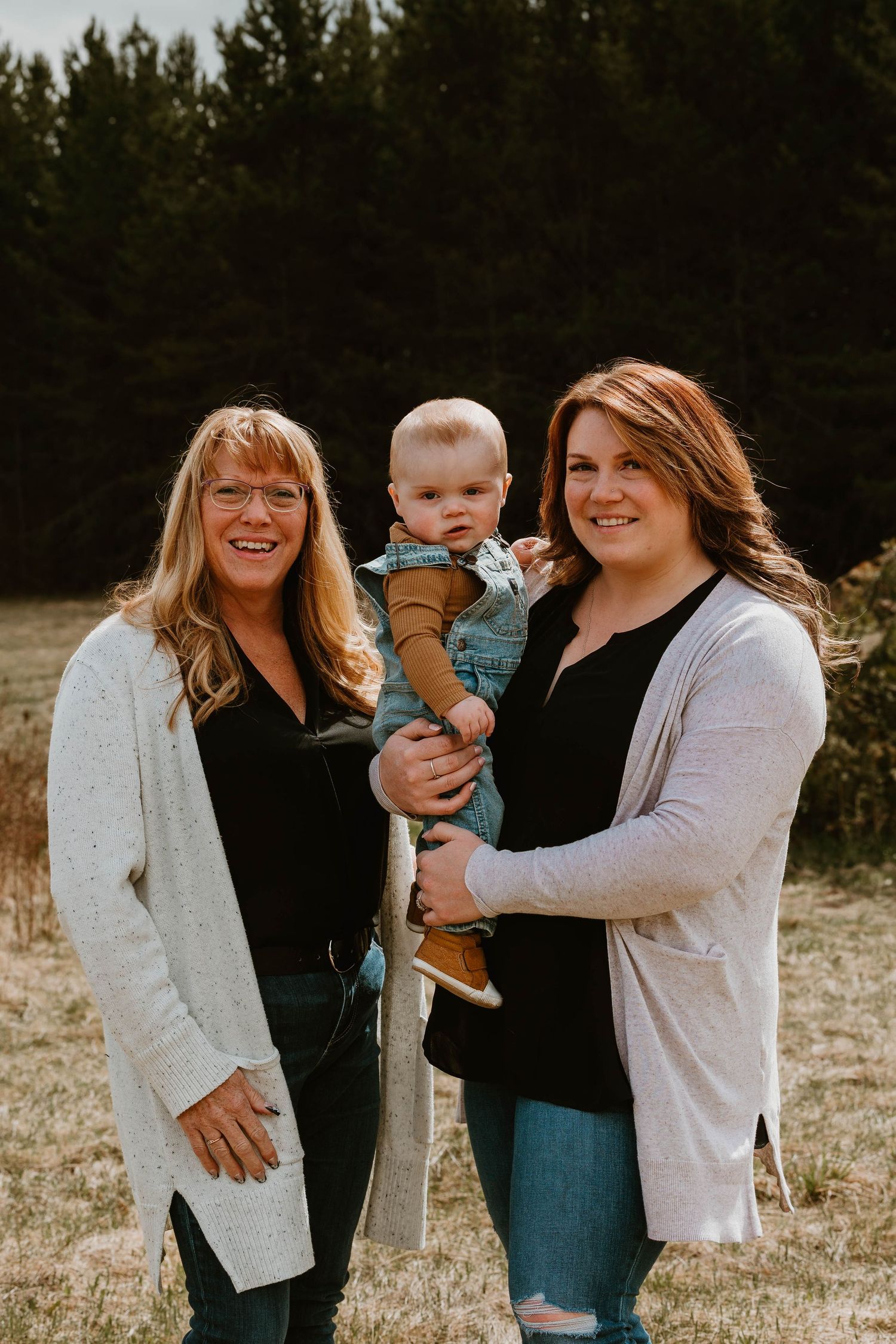 Three generations pose together outdoors against a backdrop of evergreen trees.