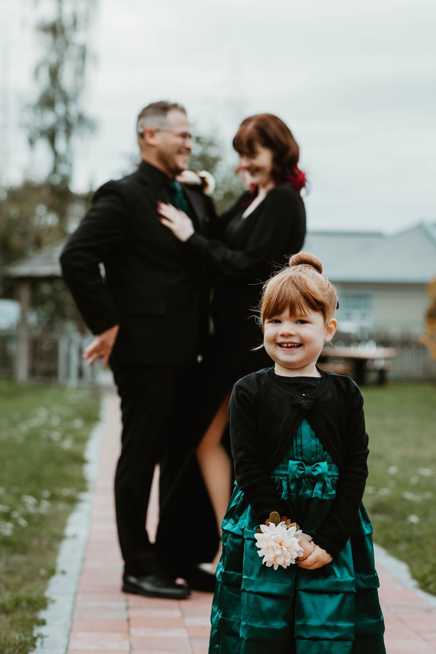 A child in an emerald dress smiles while holding flowers as two adults share a moment behind them.