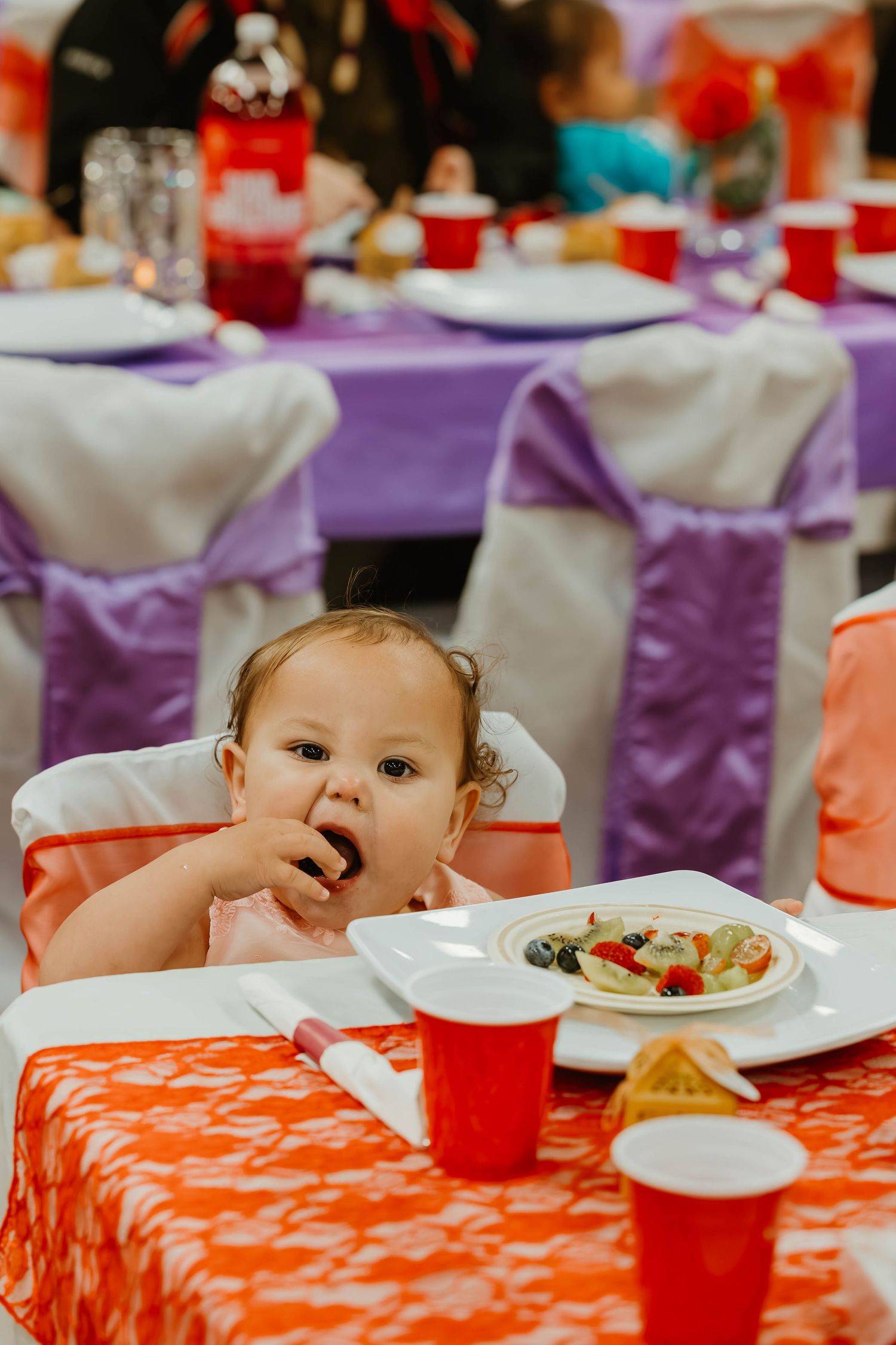 A small child eating at a decorated party table with purple tablecloths and white chair covers.