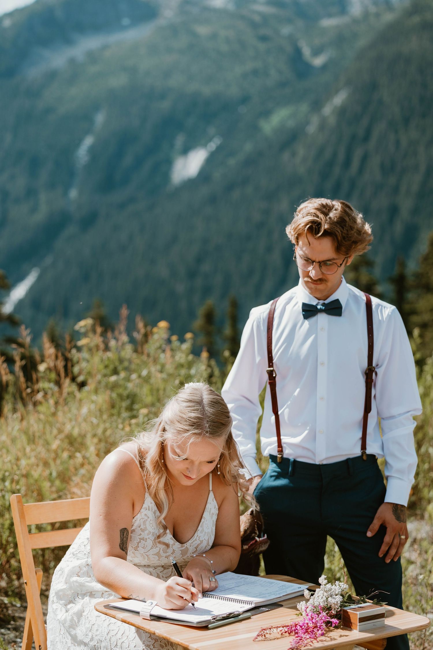 A couple signing marriage documents at a wooden table on a scenic mountain overlook.