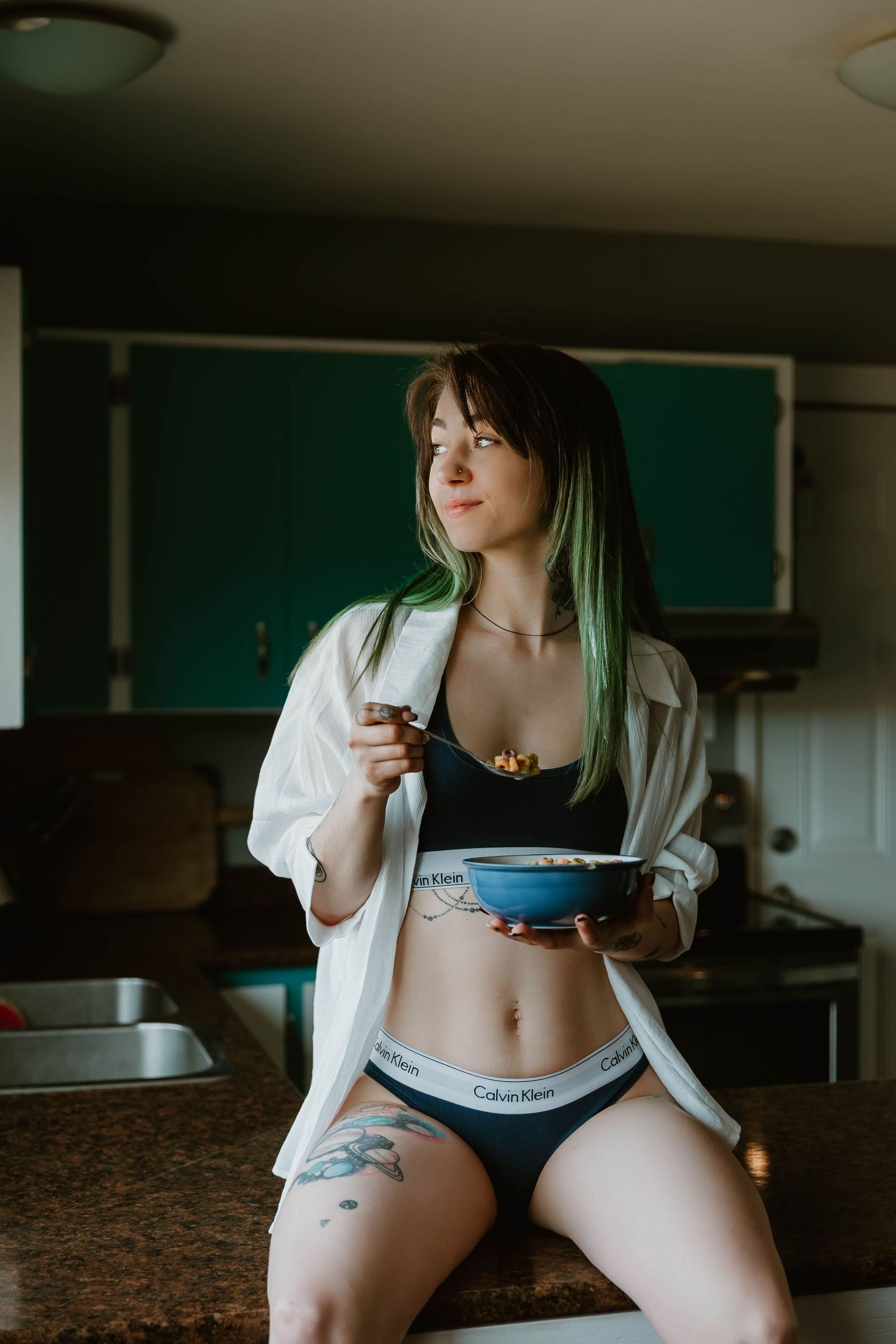 Woman with green-tinted hair wearing casual clothing sits with a cereal bowl in a kitchen setting.