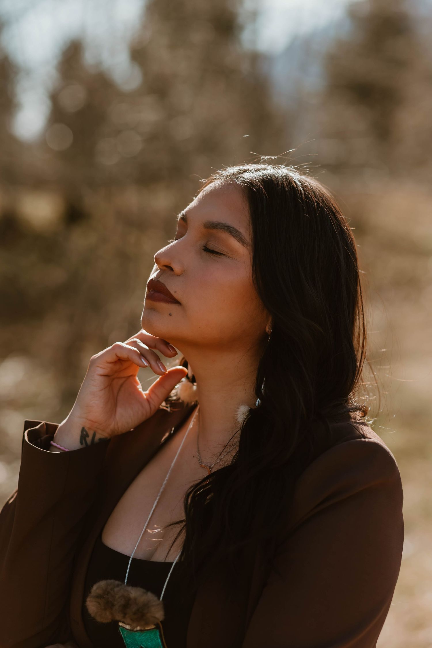 Asian woman in black blazer poses outdoors with eyes closed in warm sunlit portrait with bokeh background.