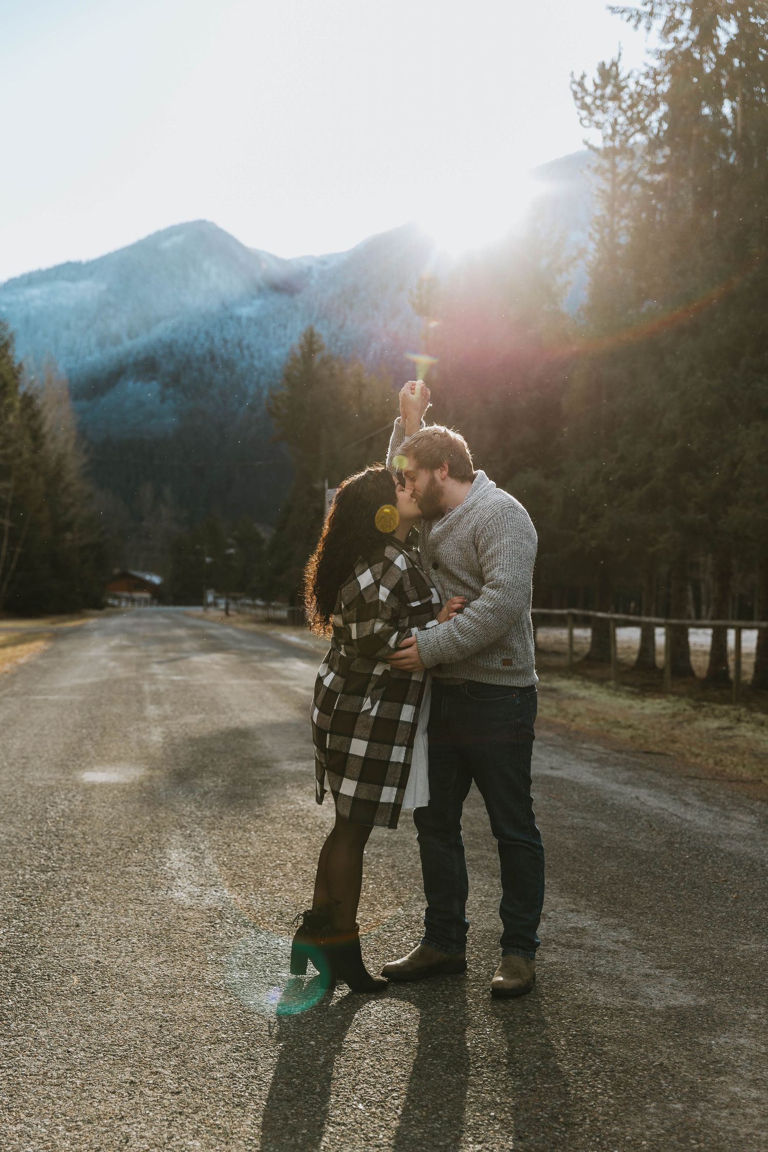 A couple embraces on a mountain road with sunlight streaming through pine trees in the background.