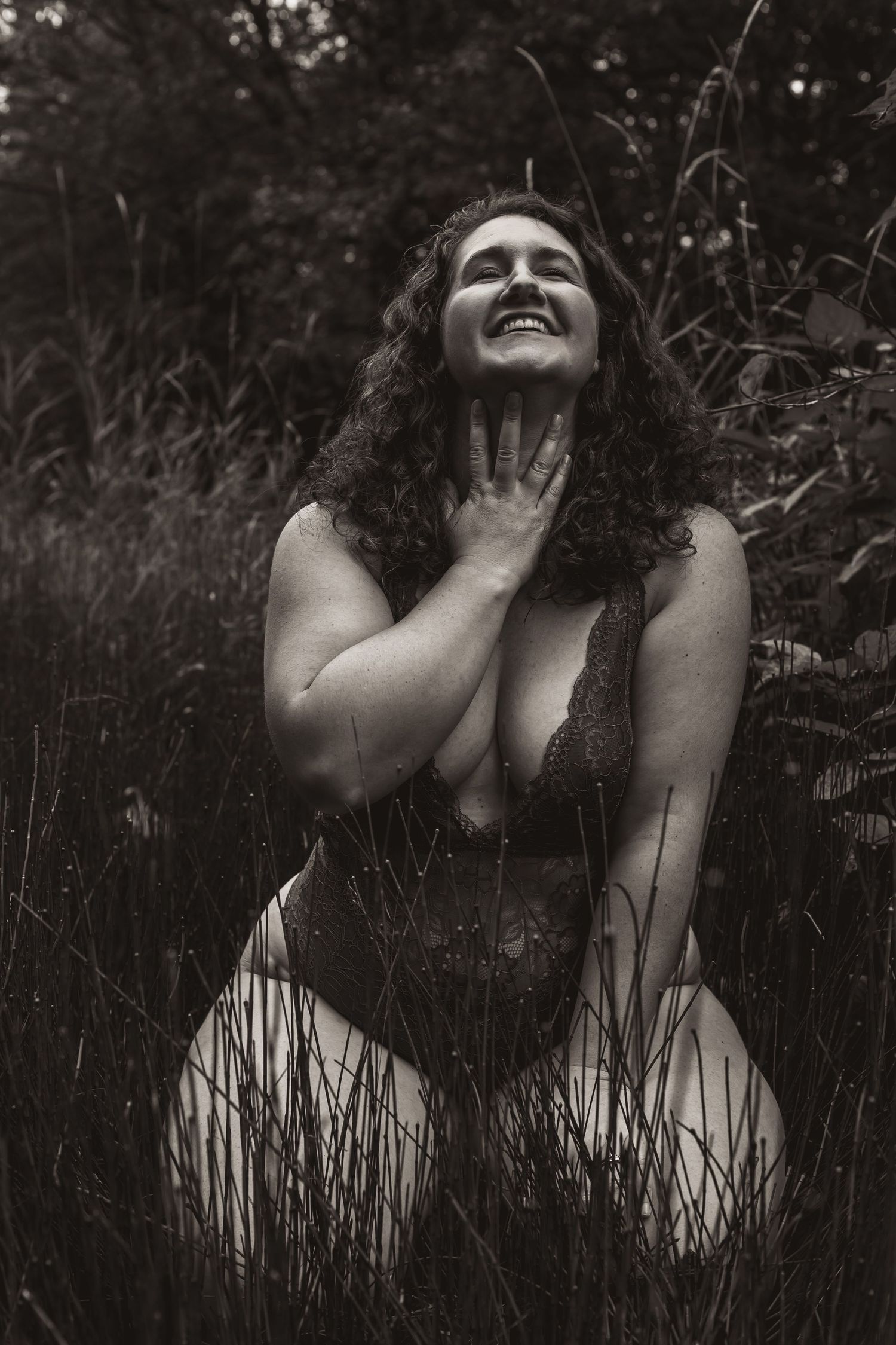 Black and white artistic portrait in tall grass shows a joyful expression and curly hair against a dark background.