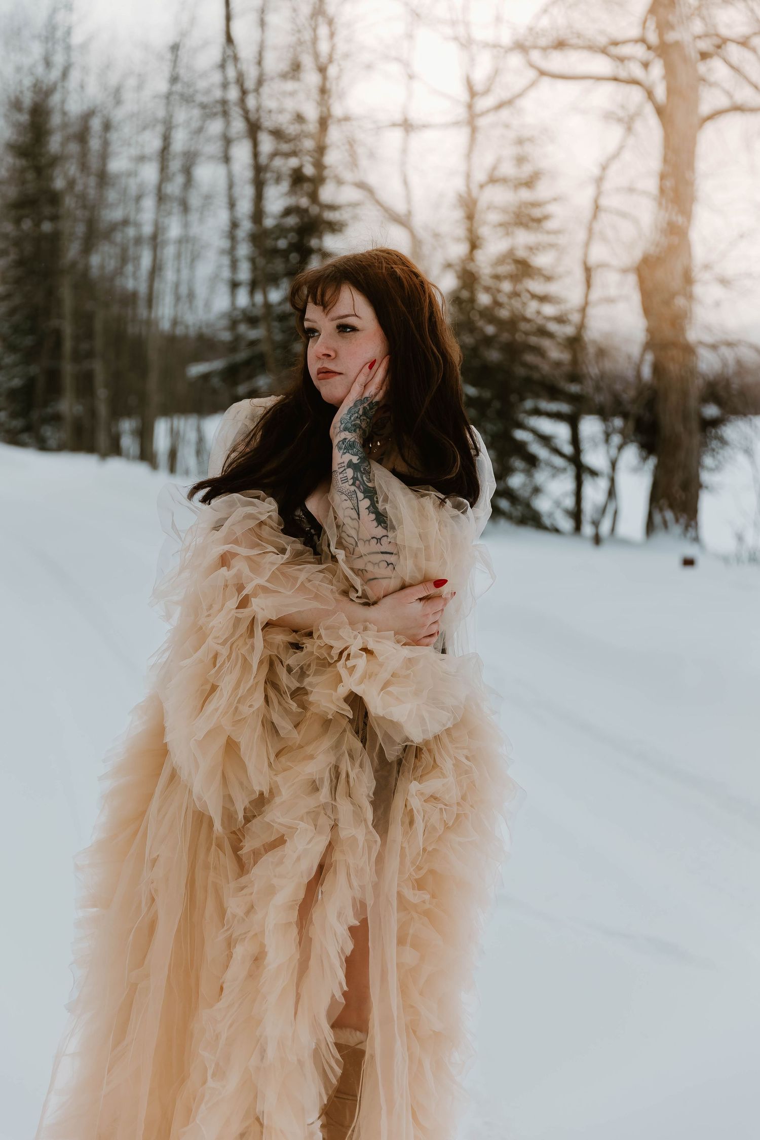 Figure wearing flowing cream tulle gown stands gracefully in snowy winter setting.