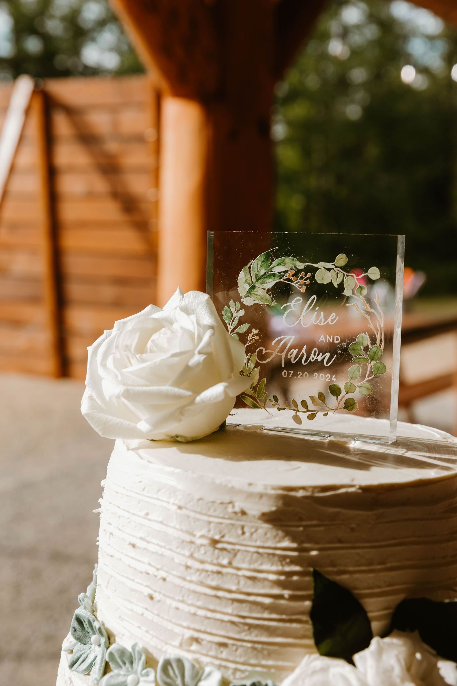 White wedding cake with textured frosting topped with rose and clear acrylic name topper.