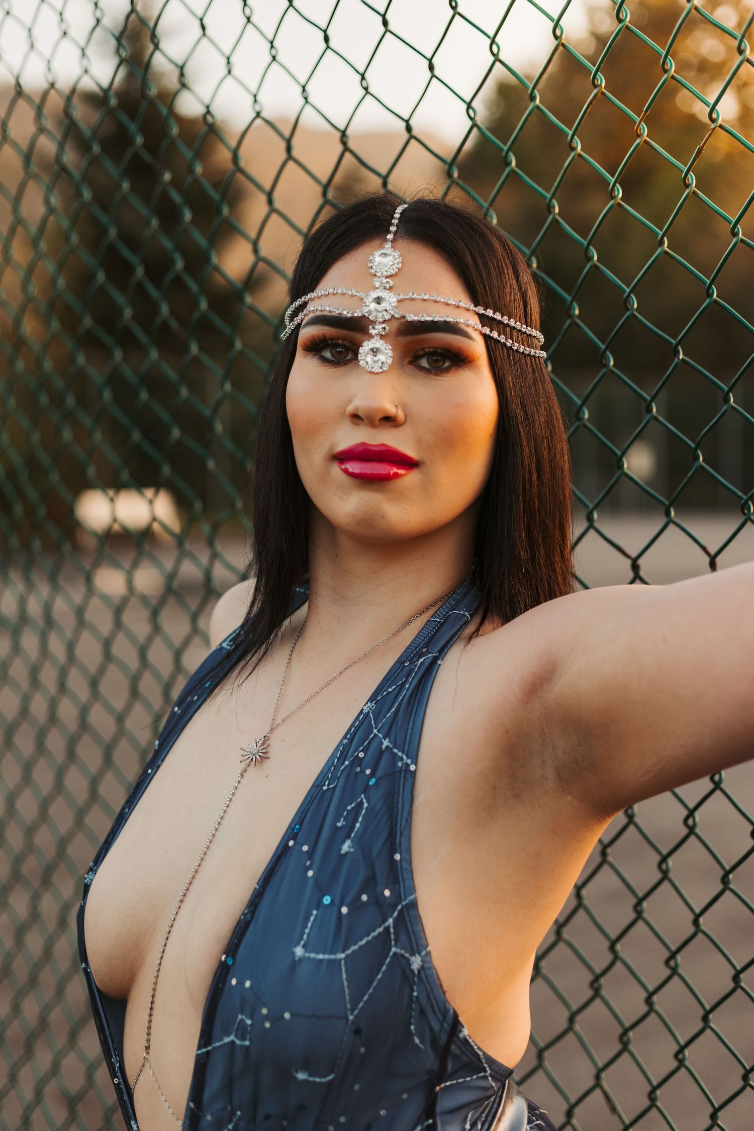 Close-up portrait with ornate headpiece and constellation print dress against chain link fence.