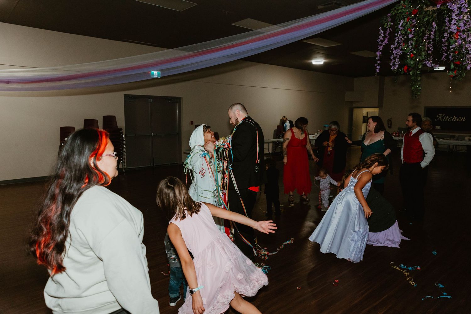 People dancing and celebrating at an indoor event with draped decorations and colored lighting.