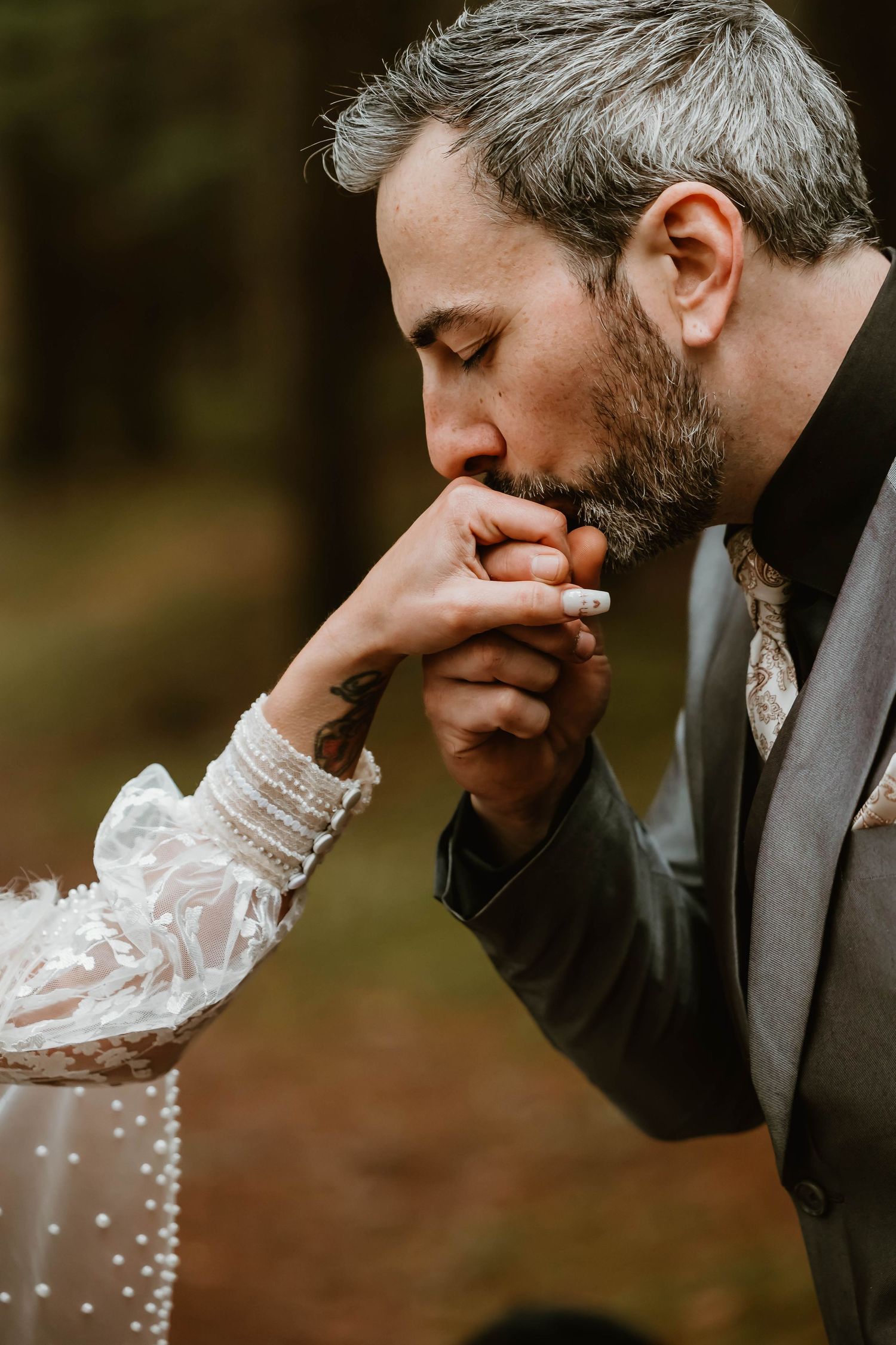 A tender wedding moment captured in the woods as the groom kisses the bride's hand, adorned in lace sleeves.