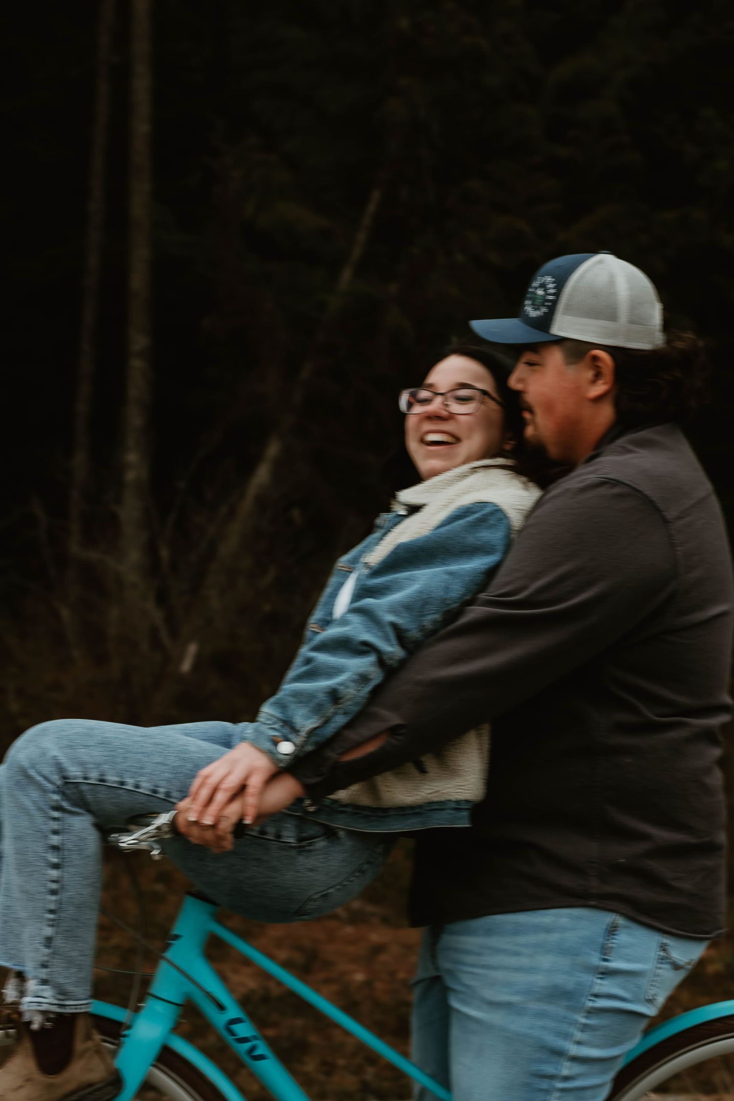 Two people share a joyful moment while riding together on a bright turquoise bicycle through a wooded area.