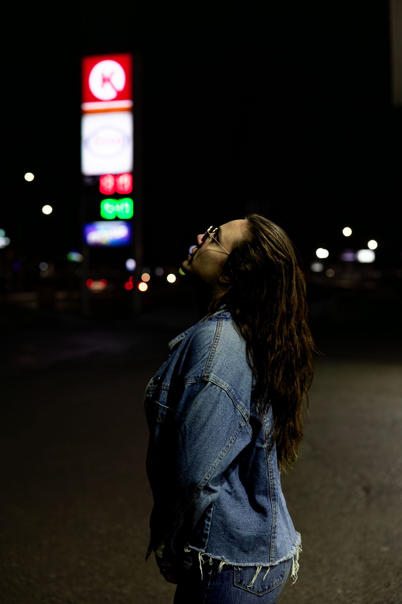 A silhouette stands in a dark parking lot at night with illuminated business signs glowing in the background.