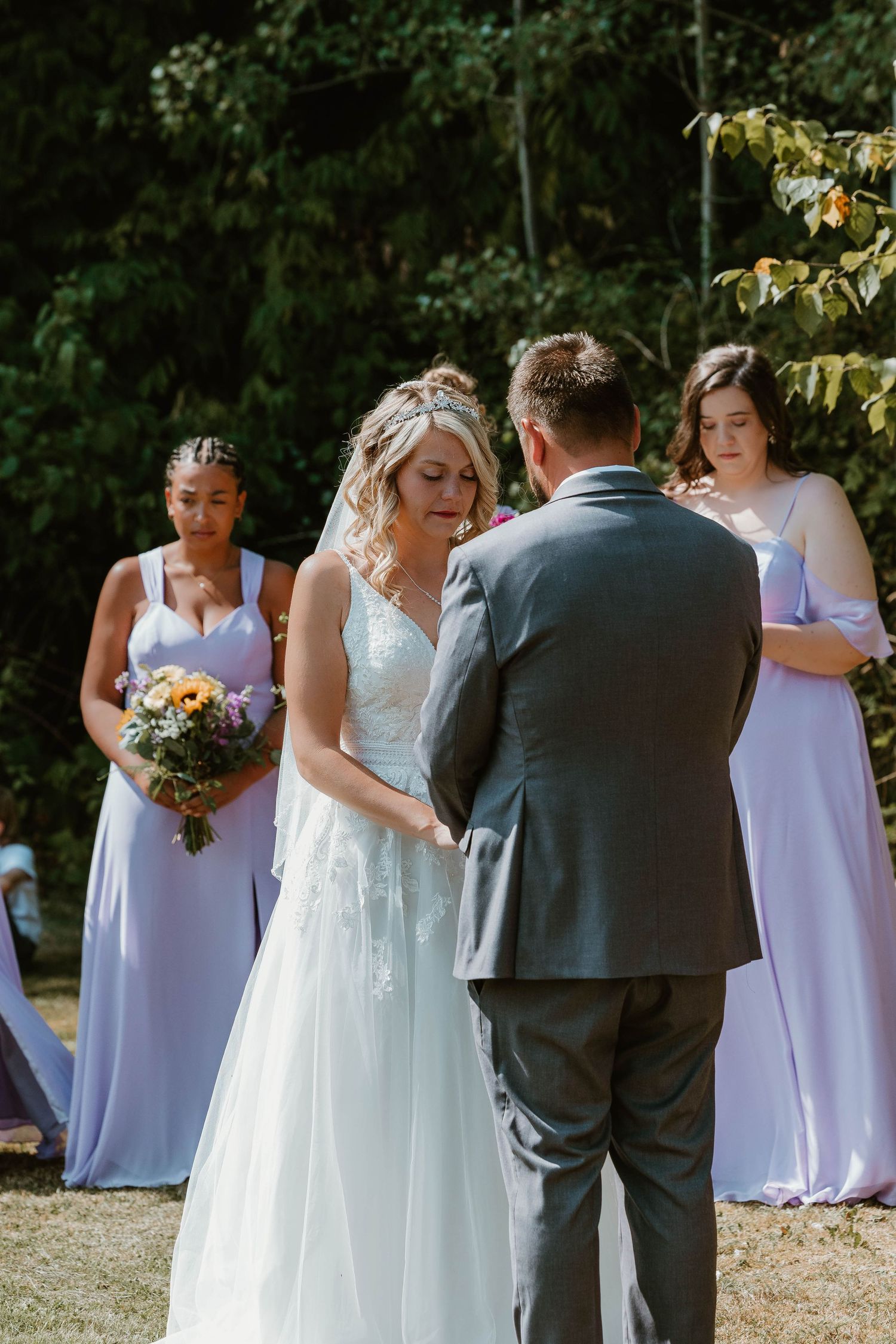 Outdoor wedding ceremony with bridesmaids in purple dresses watching the couple exchange vows.