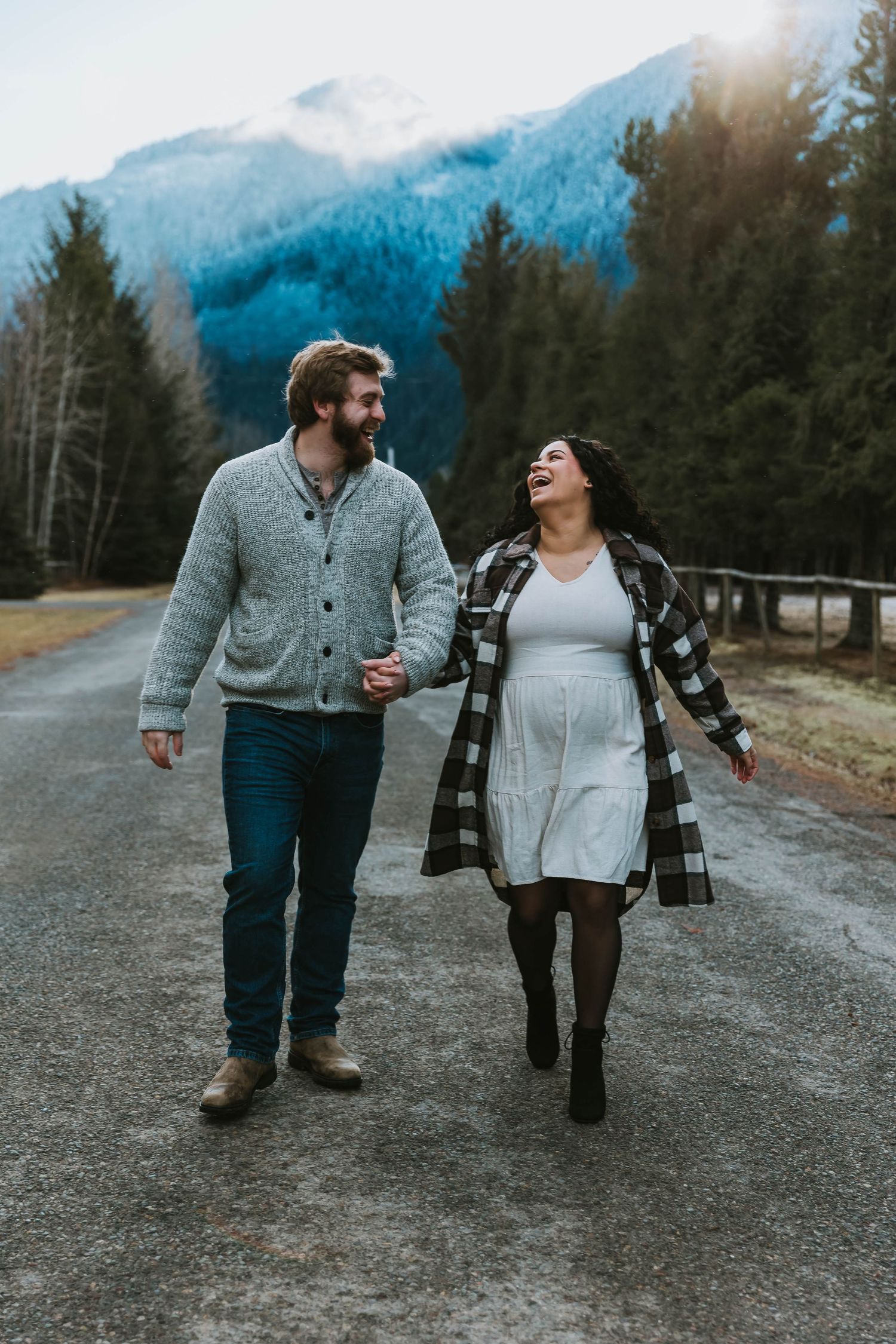 A couple walking hand in hand on a mountain road surrounded by evergreen trees and misty peaks.