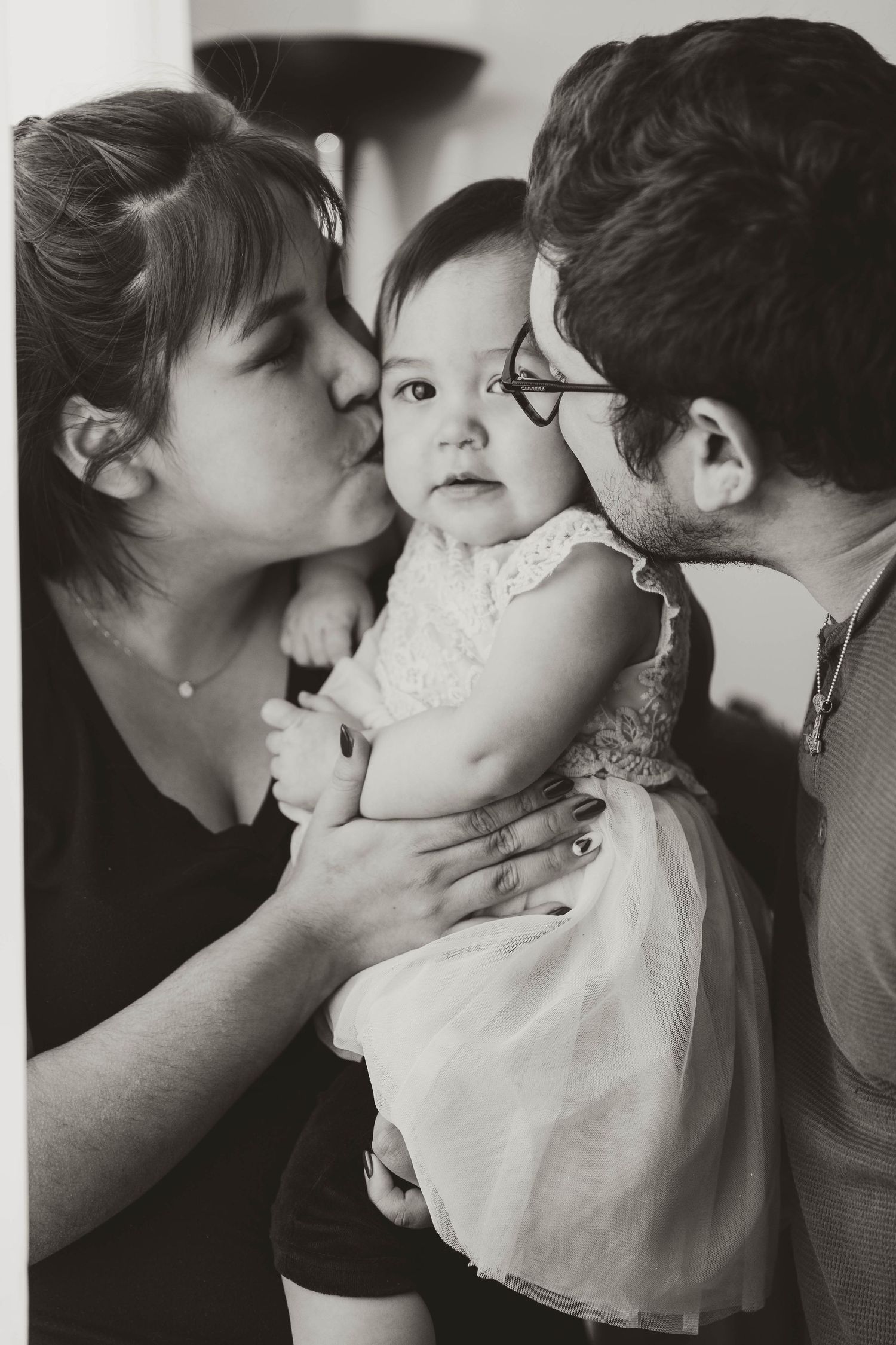Sweet black and white portrait of a baby in white dress receiving kisses from family.