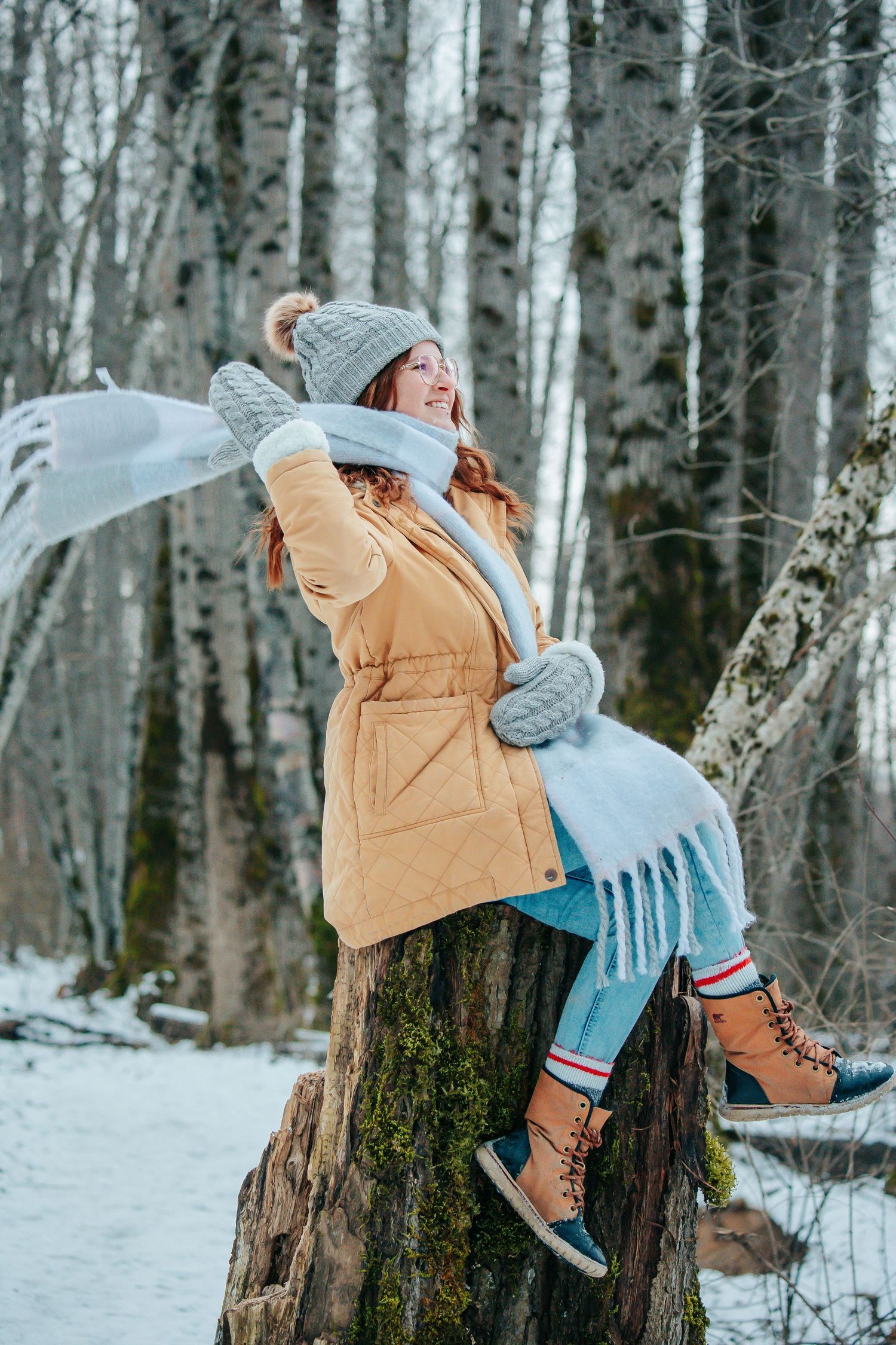 Figure in tan winter coat and knit hat poses playfully on snowy tree stump.