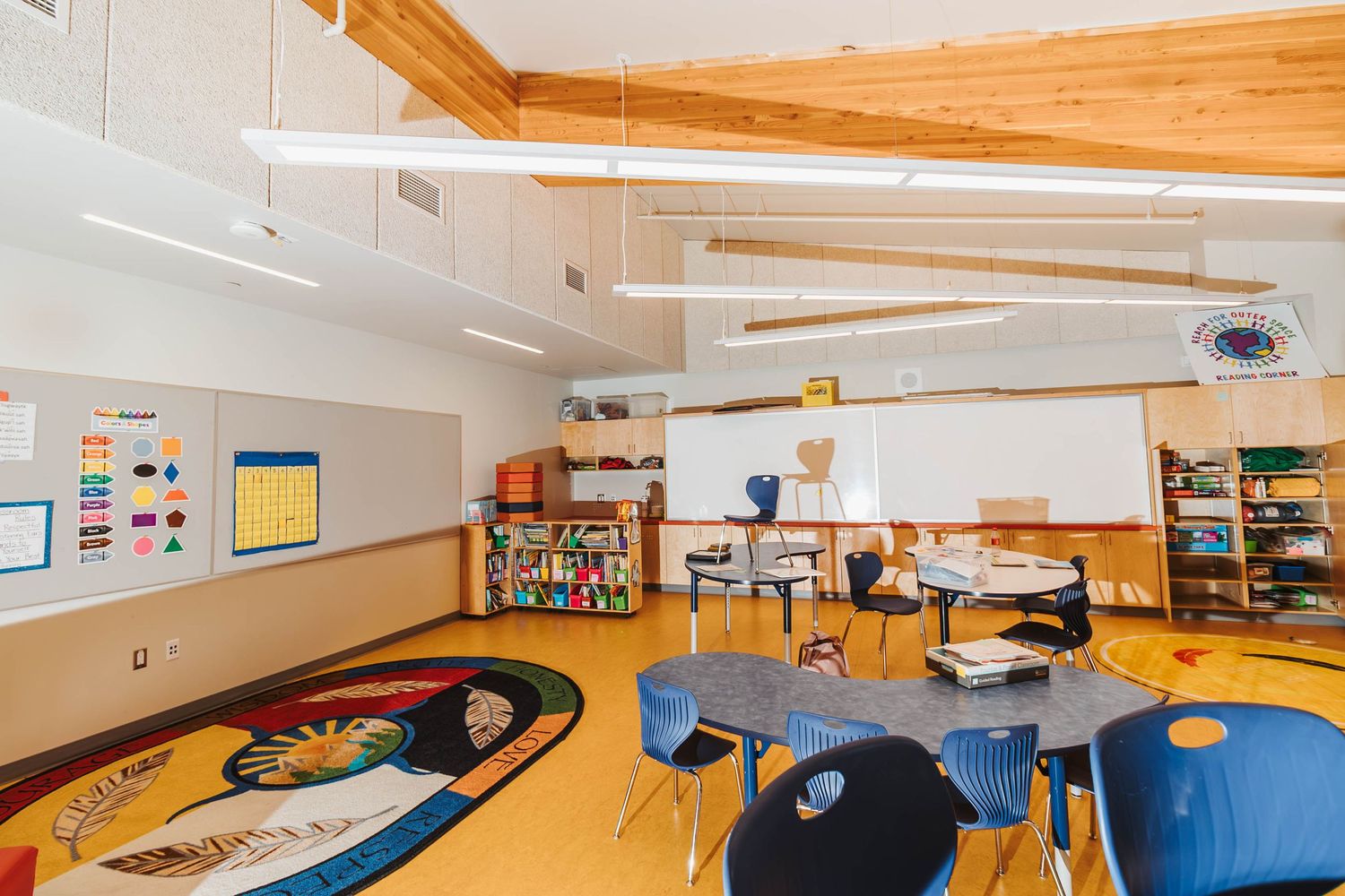 Colorful classroom with wooden beams features an area rug, blue chairs, and educational materials on walls.