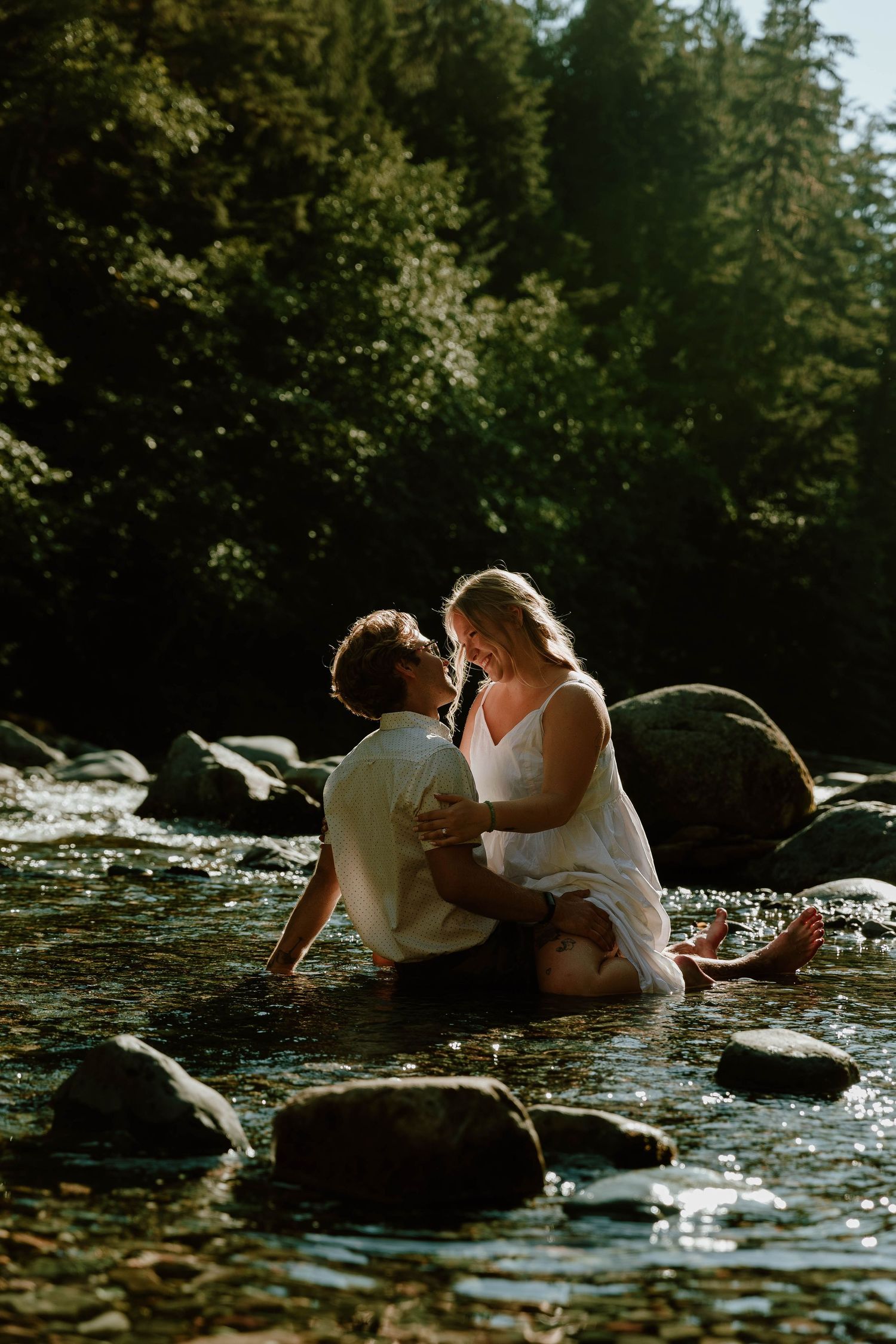 Two people embrace while sitting on rocks in a scenic river surrounded by forest.