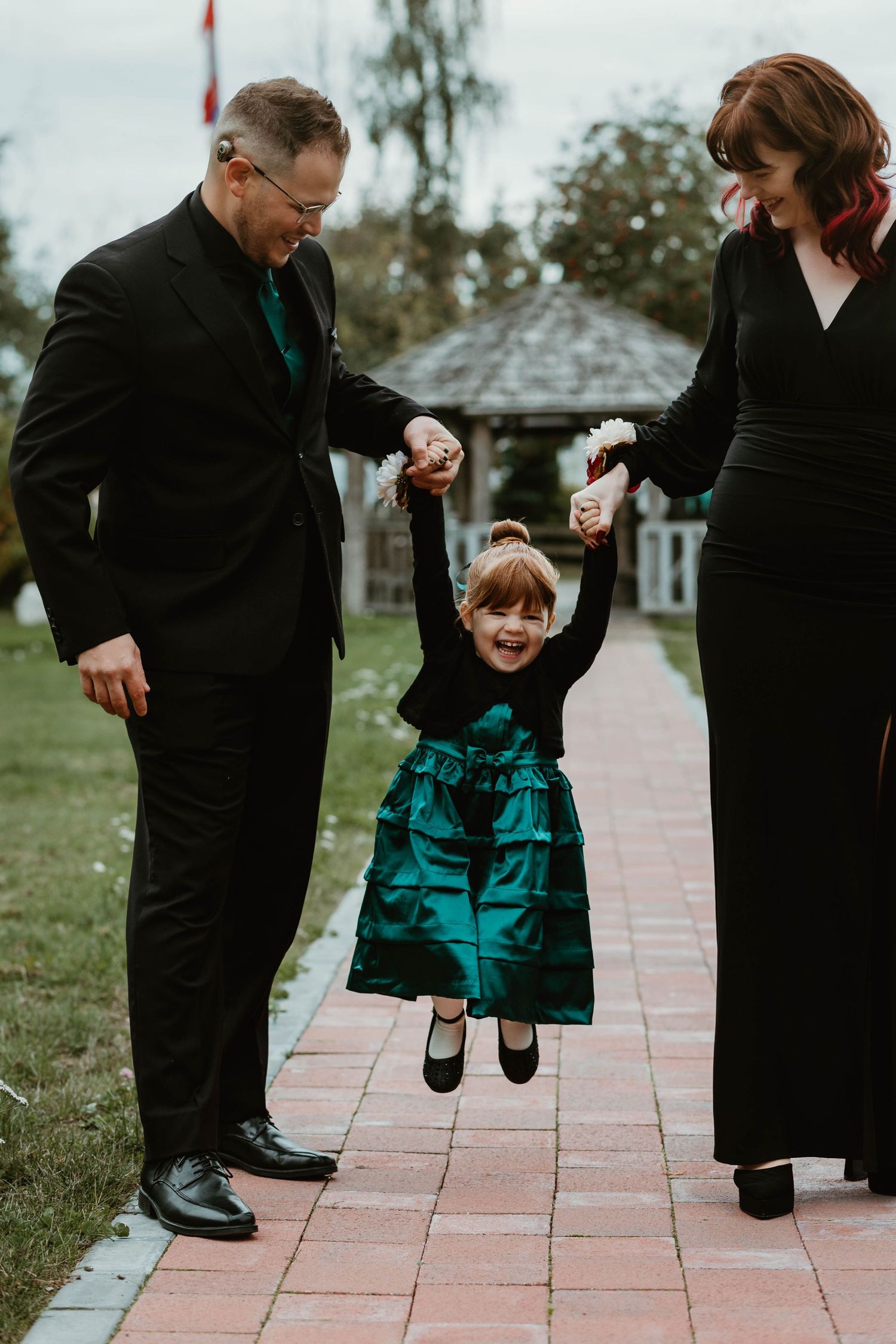 A child in teal dress swings between two adults on a brick pathway.