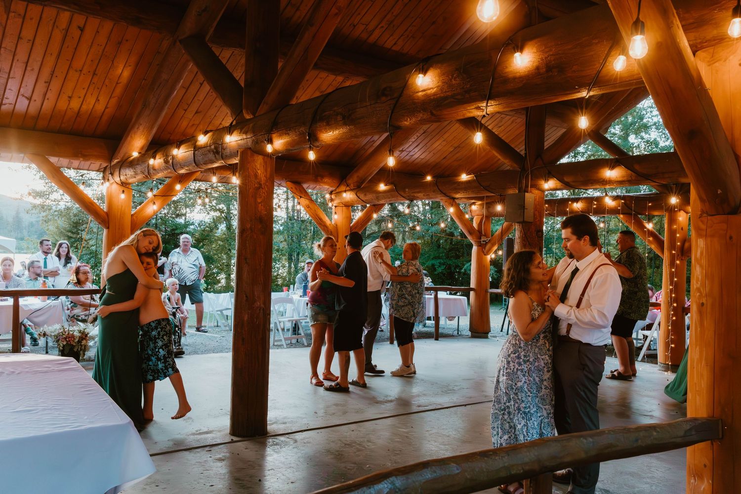 Wedding guests dancing under wooden beams and string lights at an outdoor reception venue.