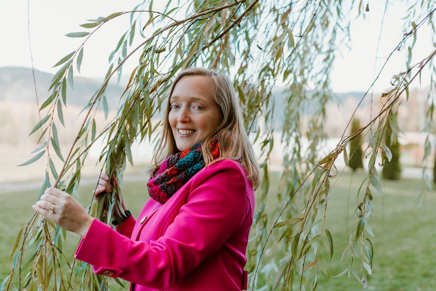 Candid laughing portrait under hanging willow branches wearing vibrant pink outerwear.
