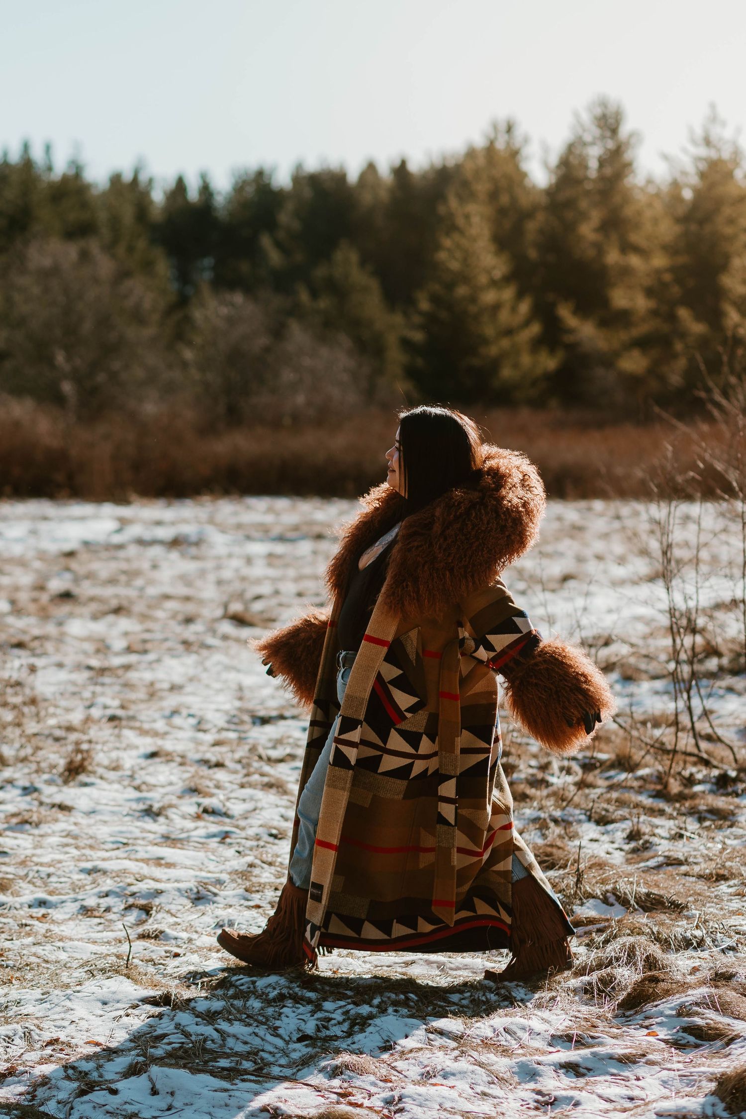Figure in a patterned winter coat with fur trim stands in snowy field at golden hour.