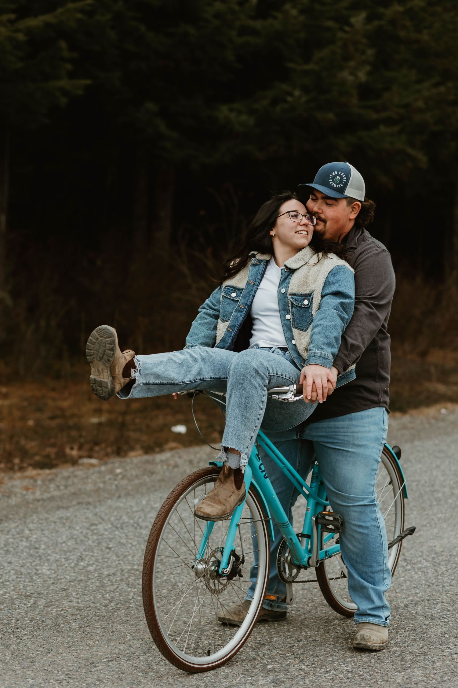 A couple shares a playful moment together on a turquoise bicycle on a rural road at dusk.
