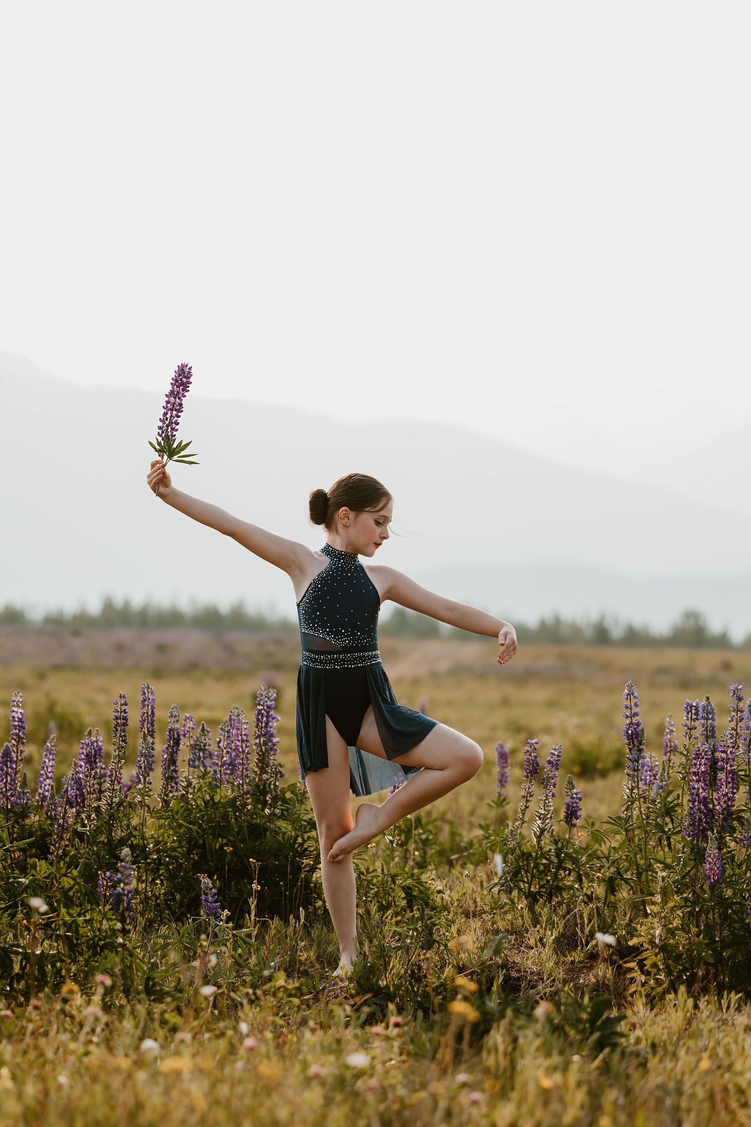 A dancer performs graceful ballet poses among purple lupine wildflowers in a mountain meadow at sunset.